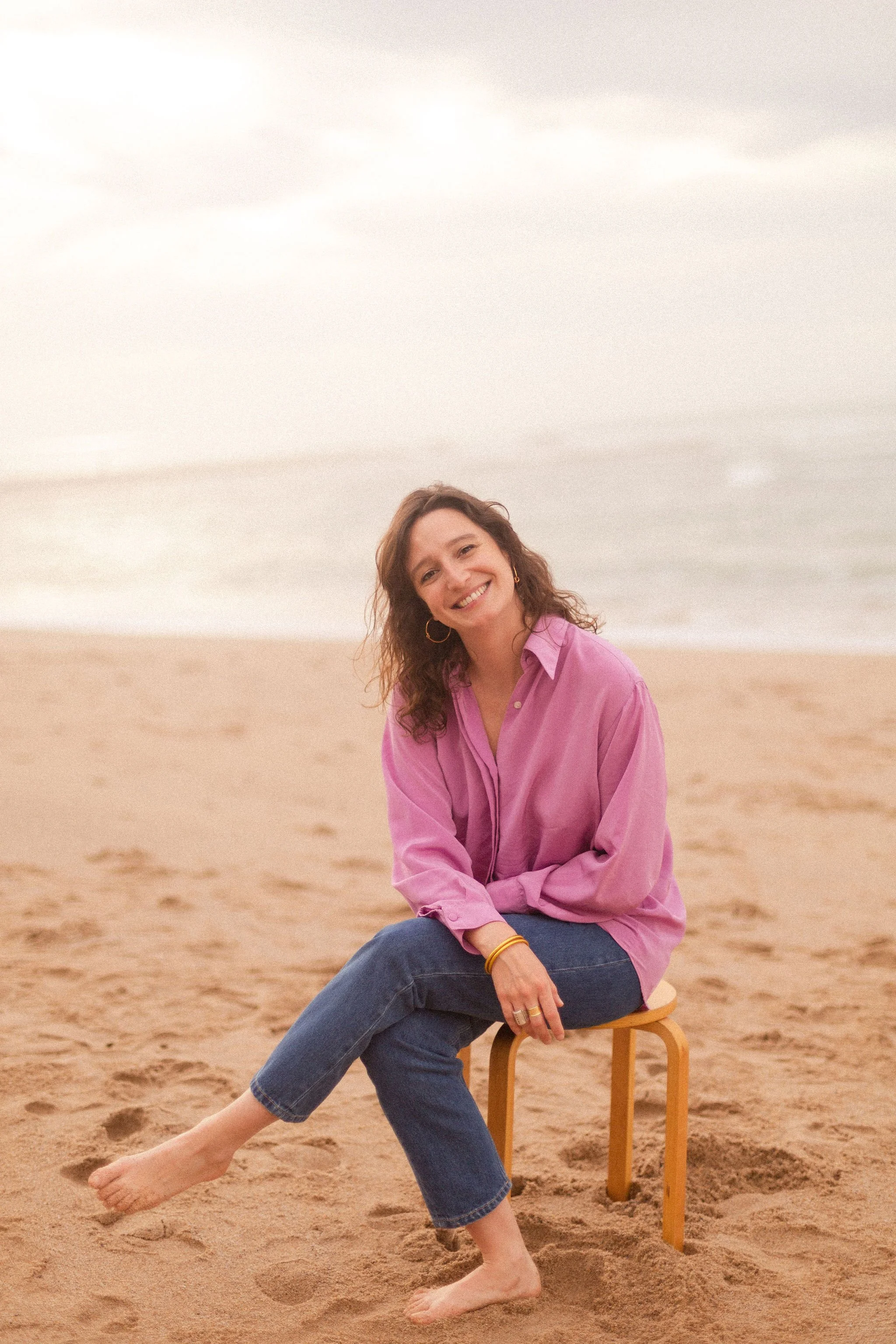 Une femme souriante en chemise rose, assise sur une petite chaise en bois sur la plage, pieds nus, avec la mer en arrière-plan.