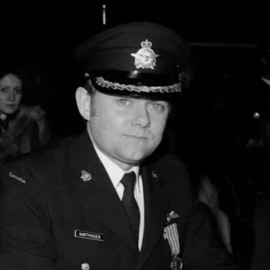 A black and white photo of T. (Terry) Hartvigsen in a Canadian military uniform, including a cap with a badge, sitting with a serious expression. A woman is visible in the background.