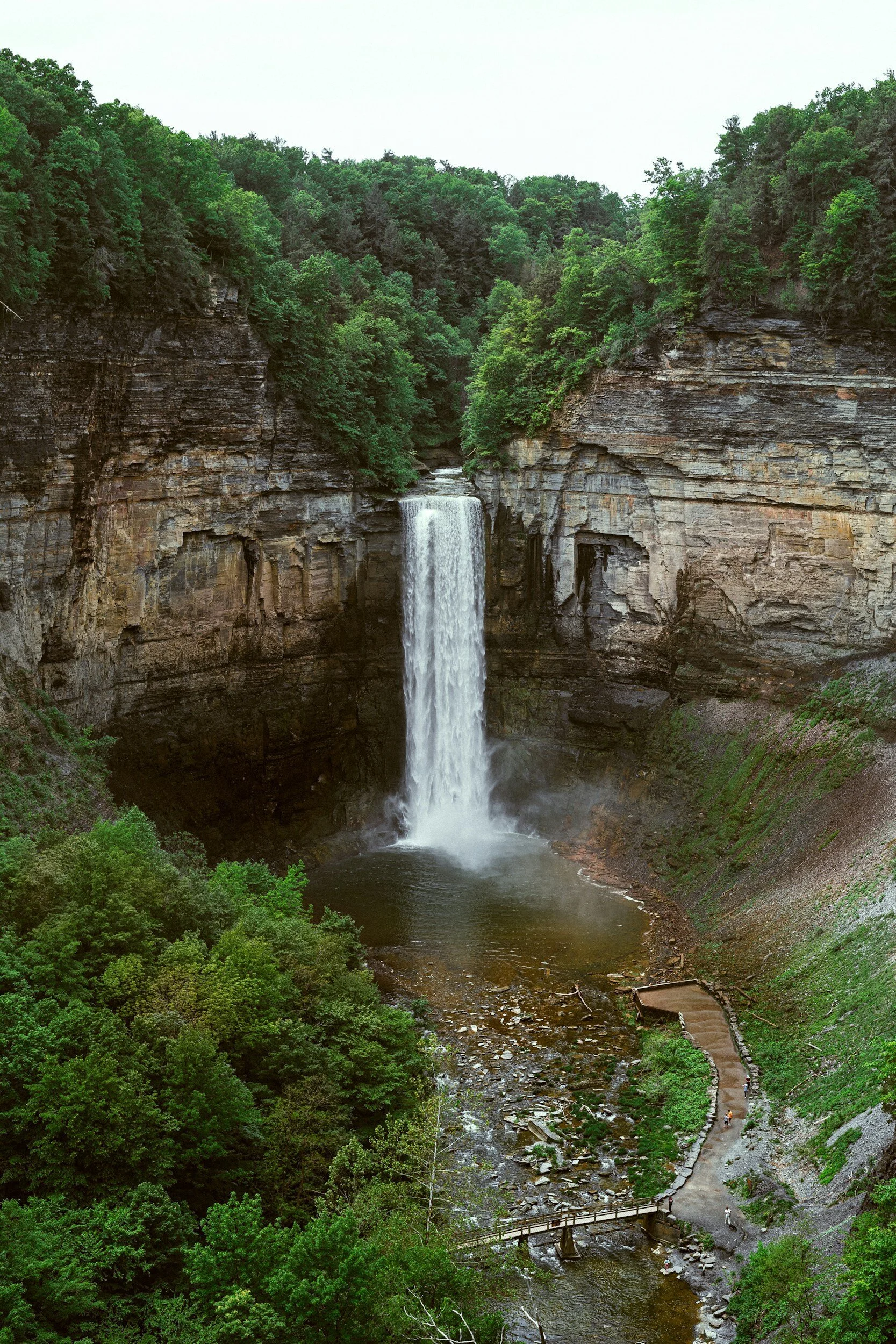 A tall waterfall cascading over a cliff into a pool below, surrounded by lush green trees and a winding pathway along the river.