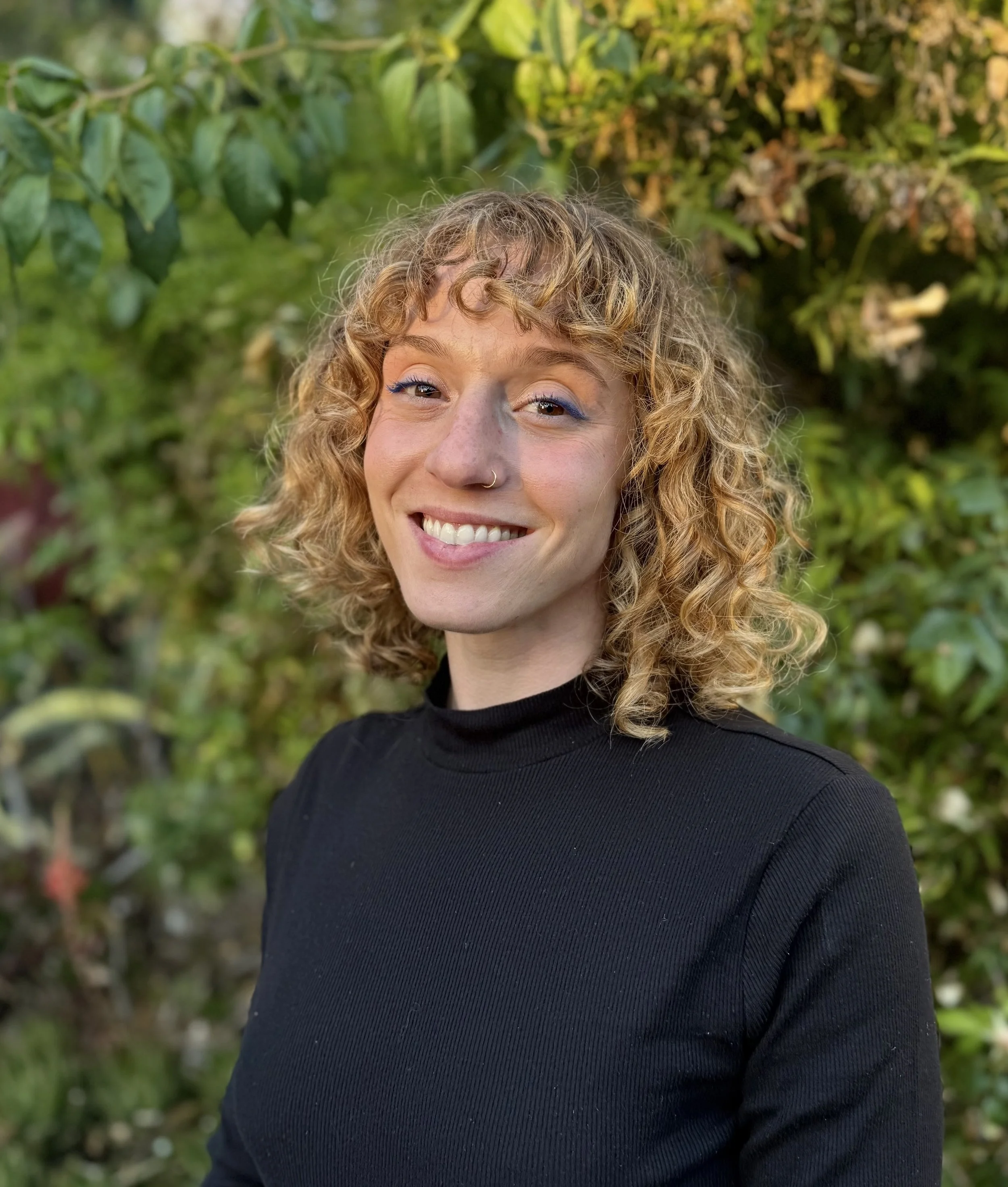 Headshot of California therapist Meli Sobin, APCC, AMFT, a woman with curly light hair with a welcoming smile, in front of greenery background