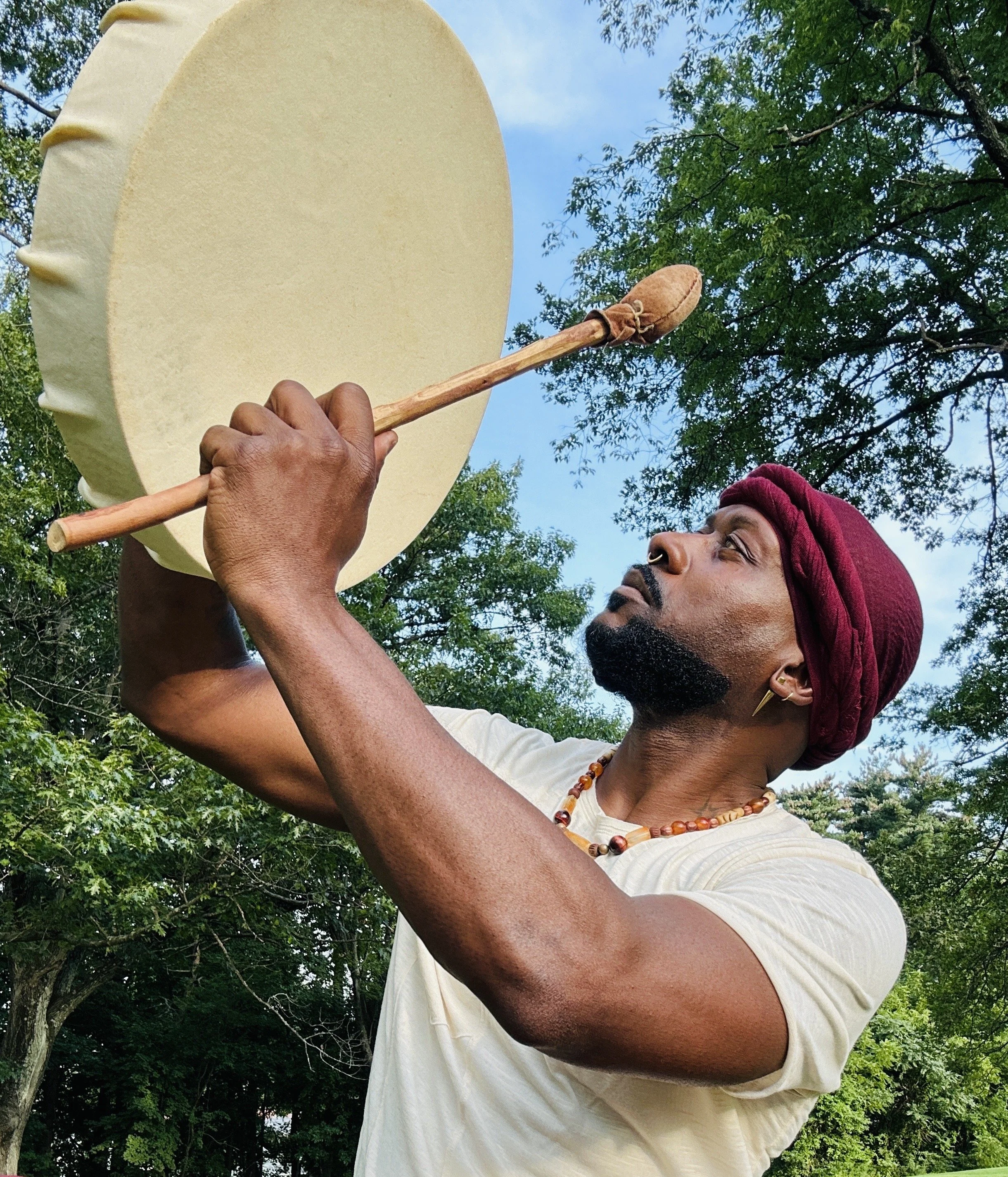 A man with a dark beard and mustache wearing a maroon head wrap and a beaded necklace, holding a large drum and a drumstick outdoors with trees and a blue sky in the background.