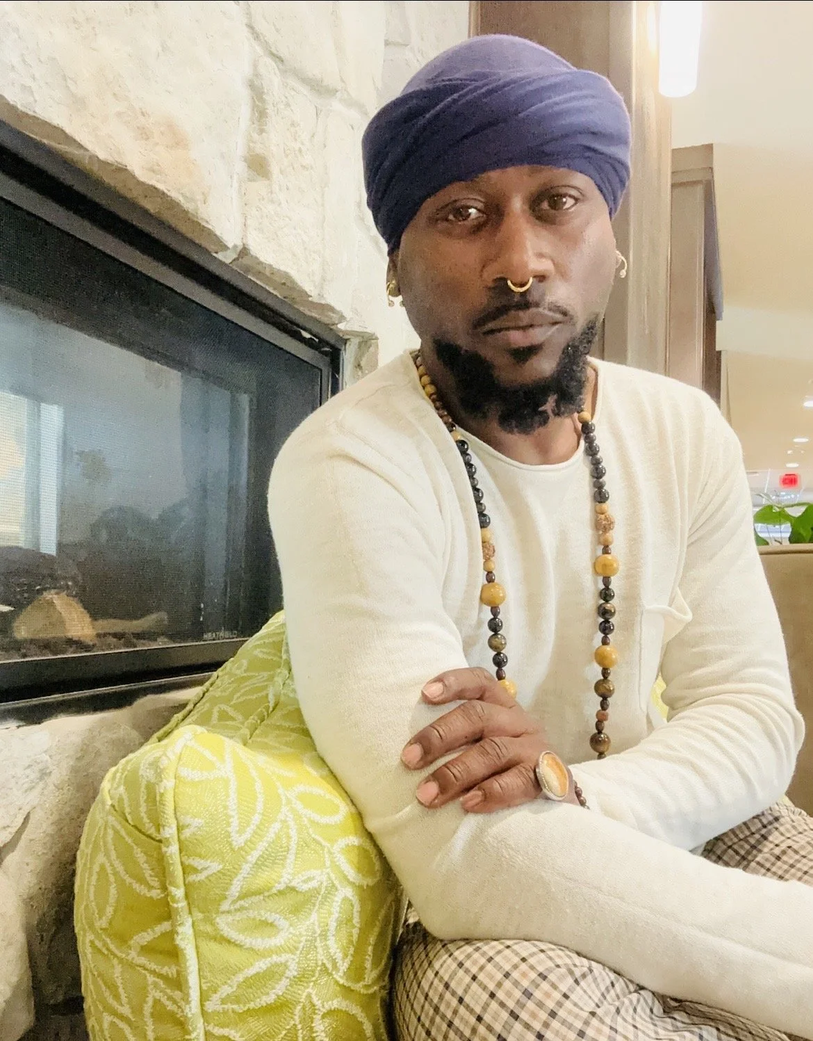 A man with a turban, gold septum ring, earrings, and beaded necklace sitting next to a fireplace with his arms crossed.