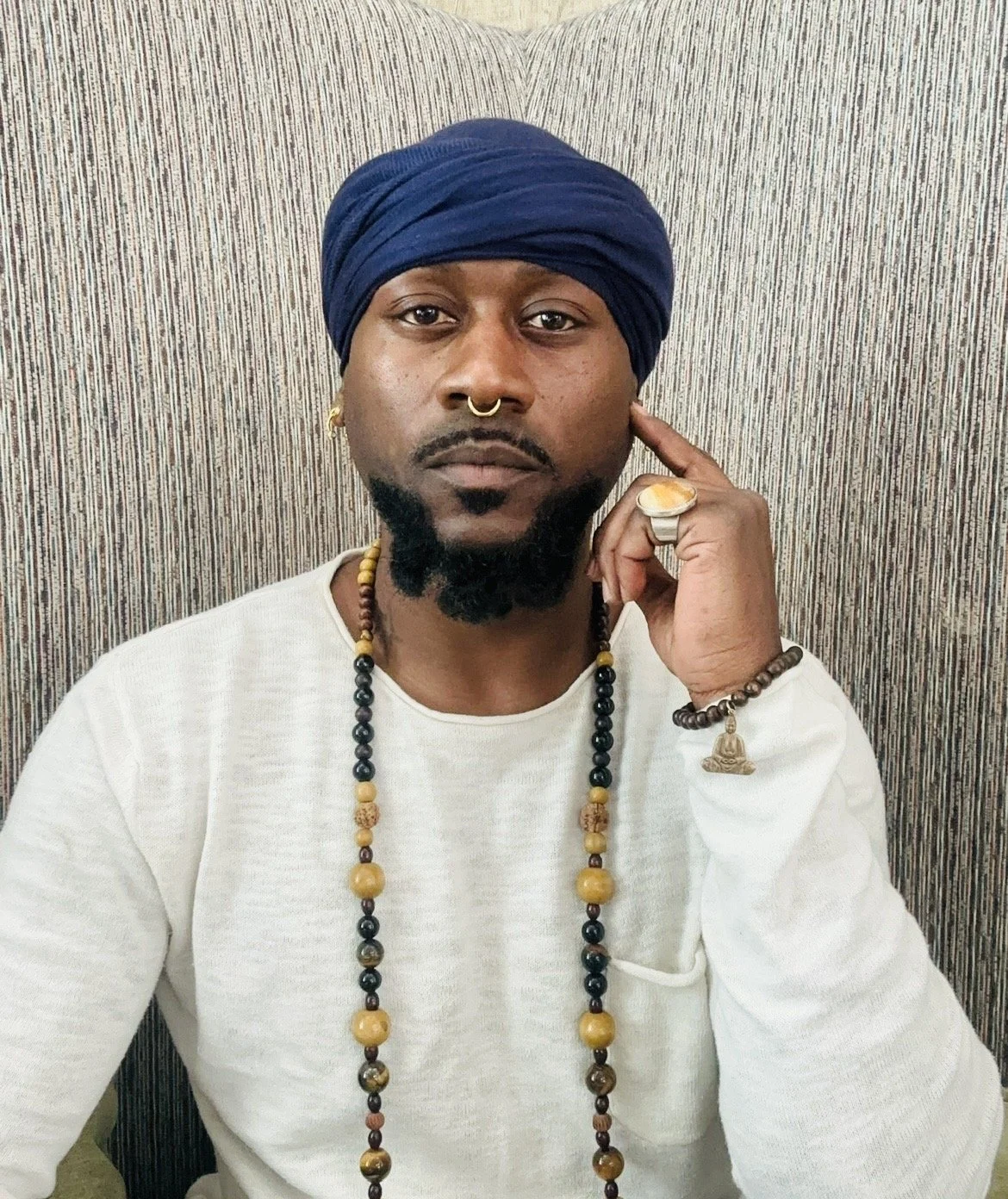 A man with a dark beard and mustache, wearing a blue turban, a septum ring, and leather beaded necklaces, sitting against a striped background, touching his temple with his right hand, wearing rings and bracelets, and a white long sleeve shirt.