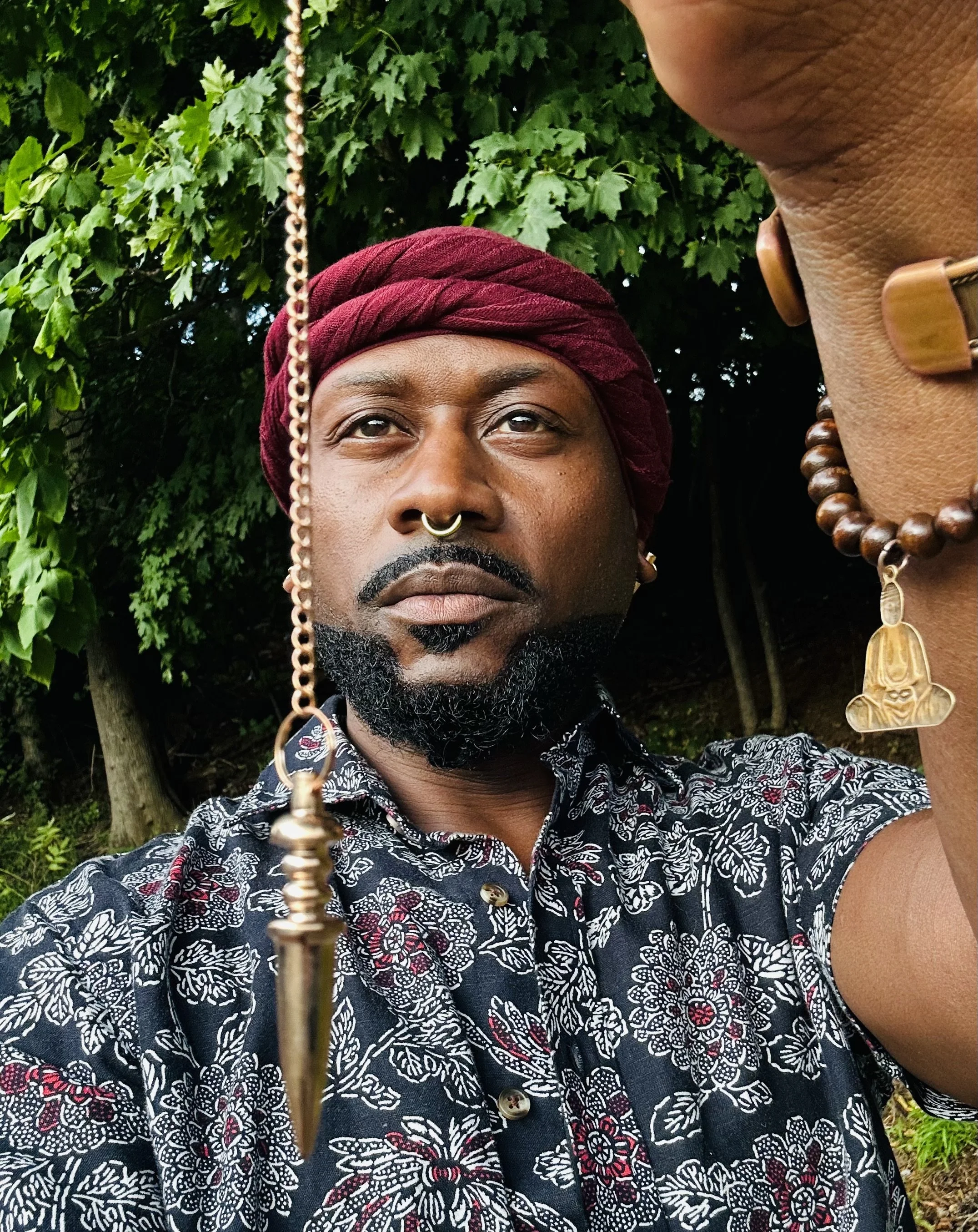 A man with medium brown skin, a beard, mustache, and septum piercing, wearing a red headwrap and a patterned shirt, taking a selfie outdoors in front of green leafy trees and bushes.