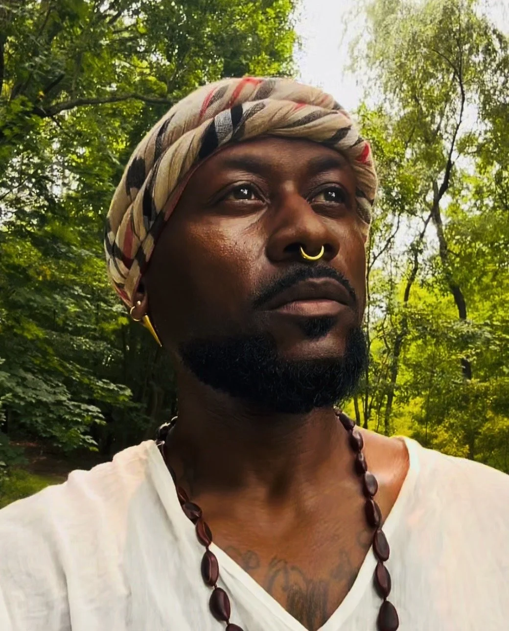 A man wearing a colorful headwrap, a septum ring, earrings, and a beaded necklace, standing outdoors among green trees.