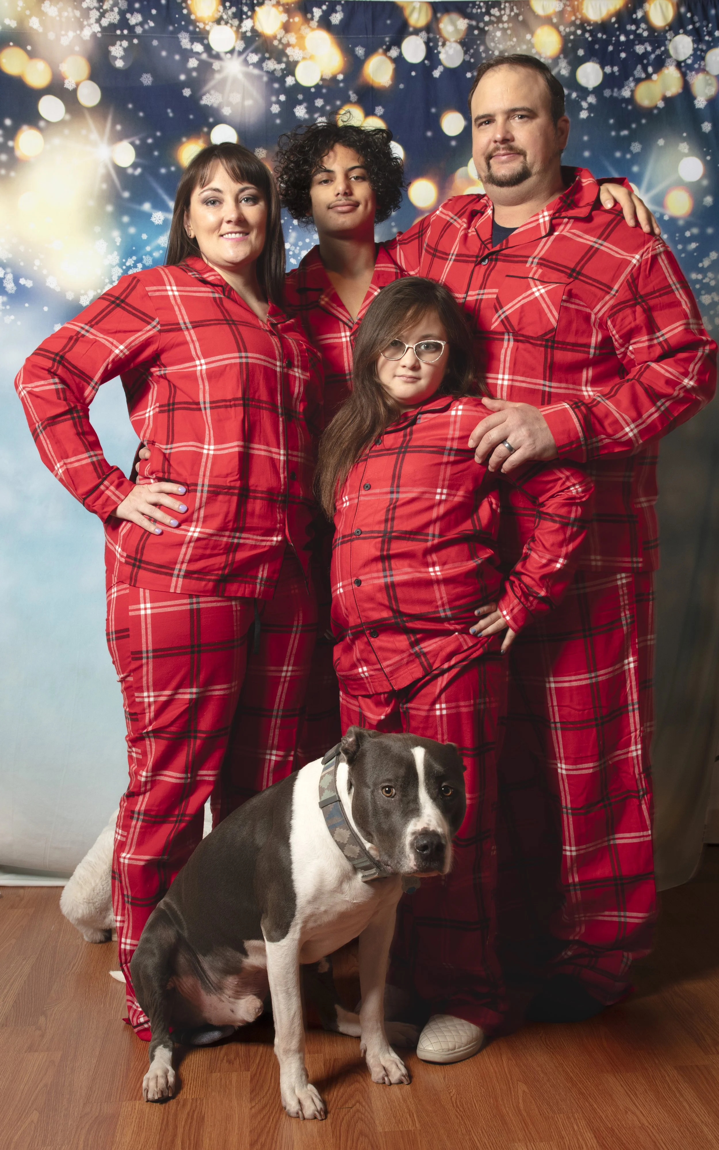A family of four in matching red plaid pajamas posed together for a holiday photo with a dog, against a festive, light-filled backdrop.