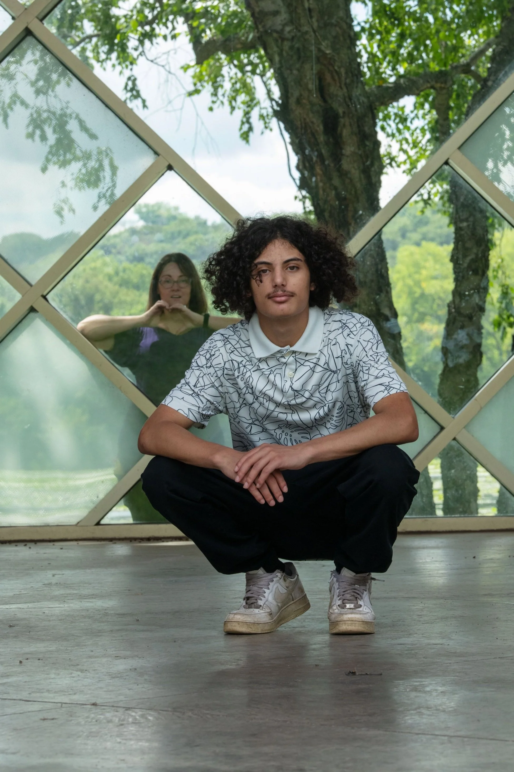Boy squatting in a pavilion in a park with mom looking in the window behind the senior
