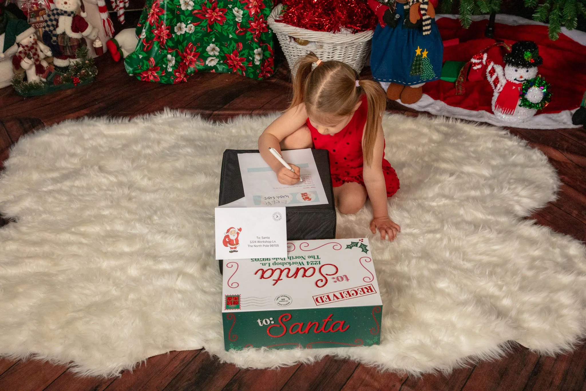 A young girl in a red dress with black polka dots is sitting on a white fluffy rug, writing a letter to Santa Claus with a pen, next to a large holiday-themed box and a letter. The background features Christmas decorations, including Santa and snowman figures, a Christmas tree, and wrapped presents.