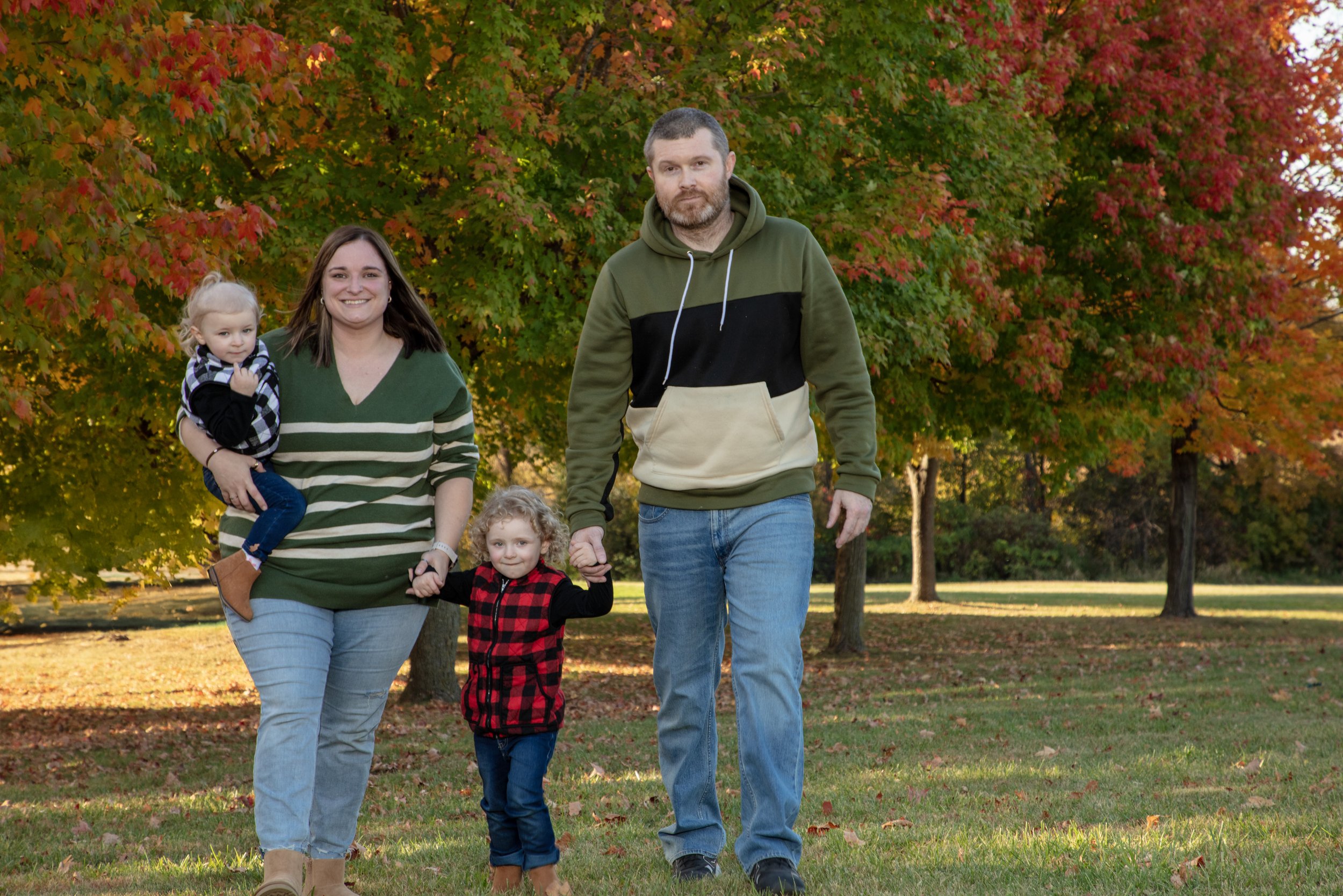 A family of four walking in a park with colorful autumn trees in the background. The woman is holding a young girl, and an older girl is holding the man's hand. They are dressed casually and smiling.