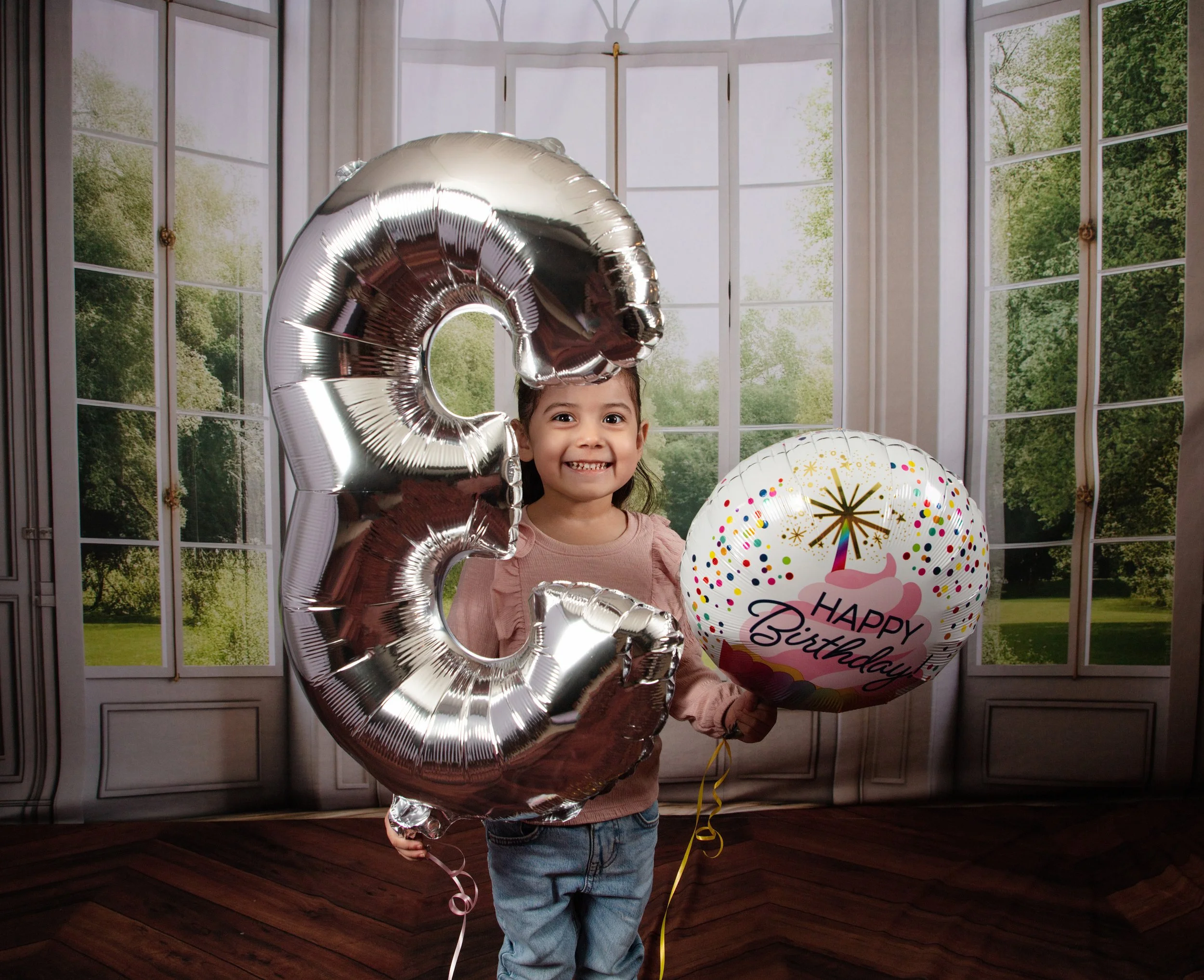 A young girl holding birthday balloons, one shaped like the letter 'G' and the other with a birthday cake and 'Happy Birthday' message, standing in front of a large window with a view of trees.
