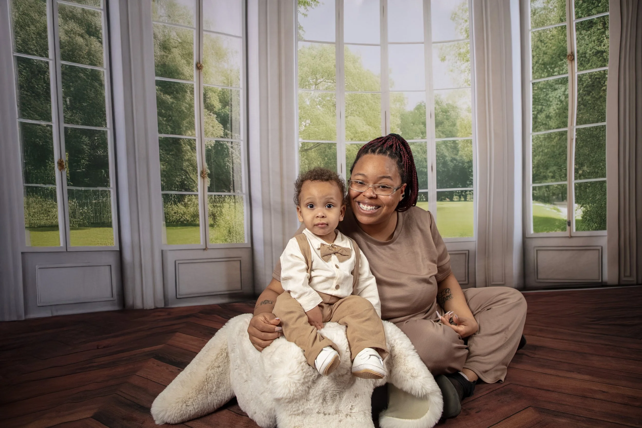 A woman and a young boy sitting on a soft white blanket in front of large windows with a view of outdoor greenery, smiling at the camera.