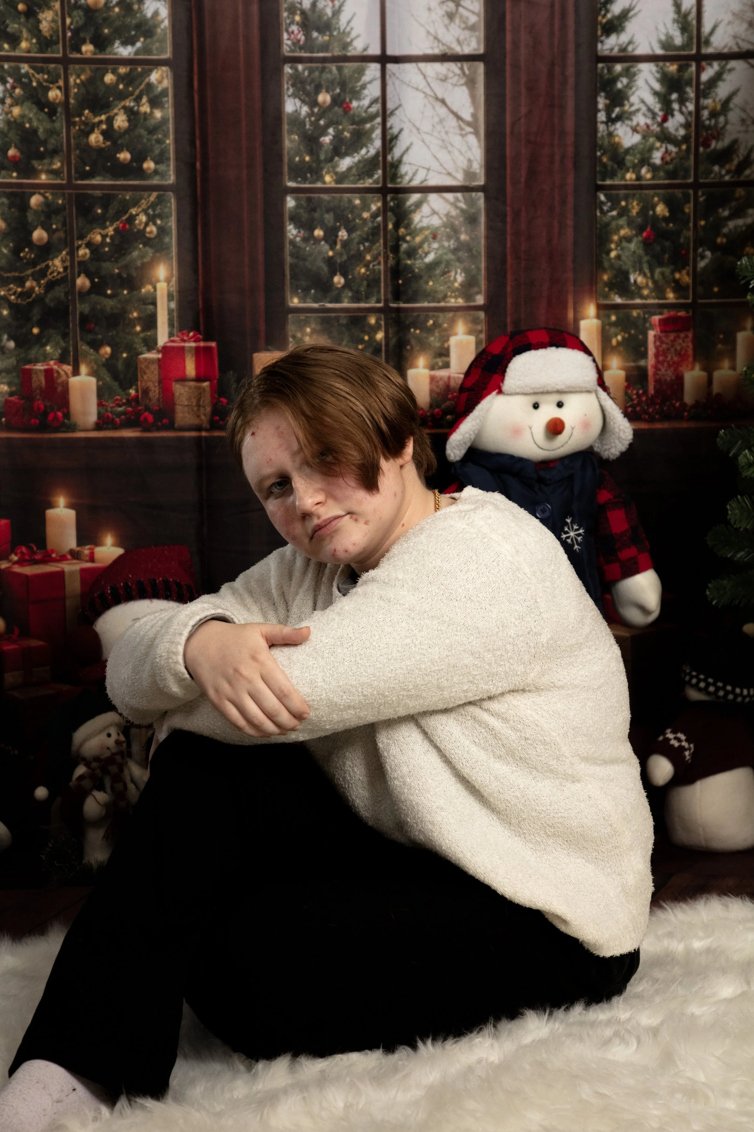 A young person with acne and brown hair sitting on a white fluffy rug in front of a Christmas-themed backdrop, surrounded by stuffed snowmen and Christmas presents.
