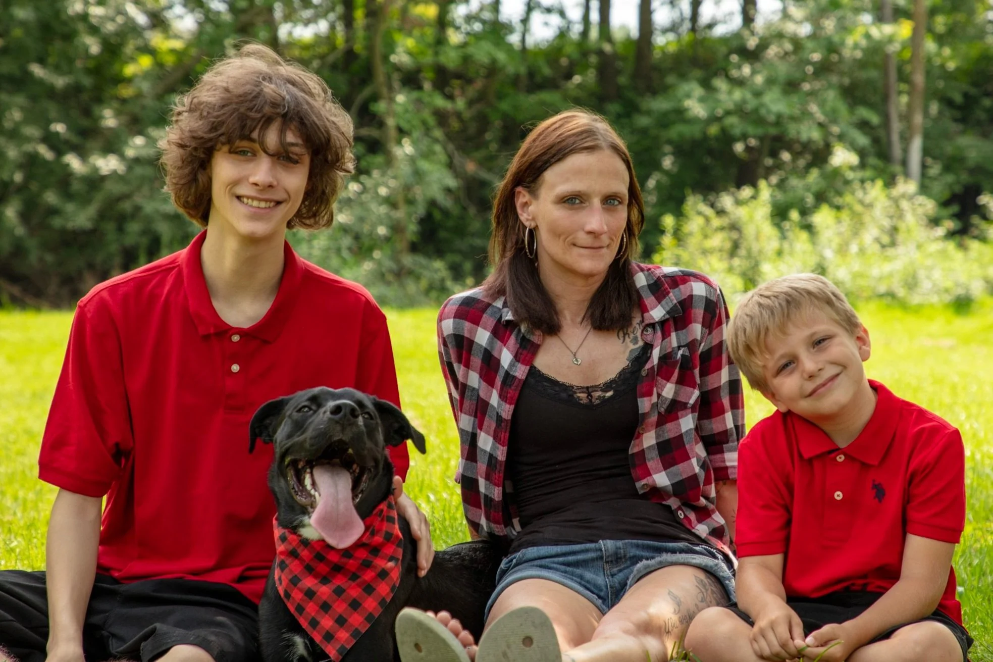 A woman and two children sitting on grass outdoors with a black dog, all dressed in red, smiling at the camera.
