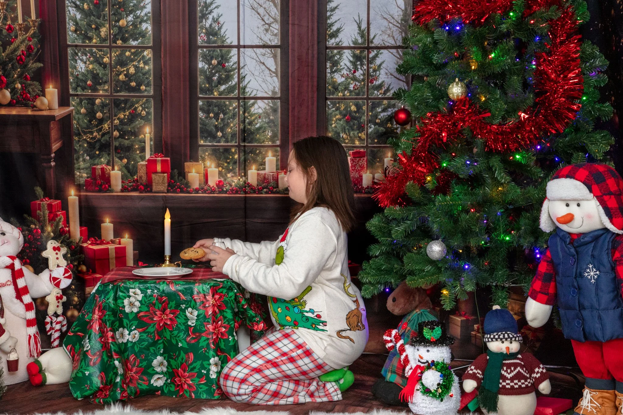 Girl kneeling at decorated Christmas table with lit candles, festive decorations, and a Christmas tree adorned with ornaments, surrounded by plush snowmen and other holiday decorations.