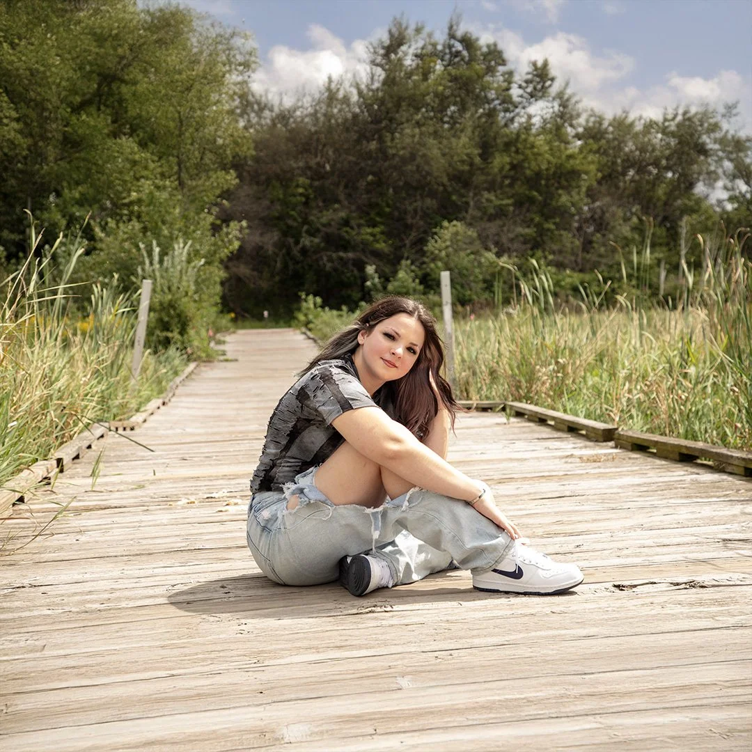 girl in the park, sitting on a wooden walk path, wearing jeans and a black and grey shirt.