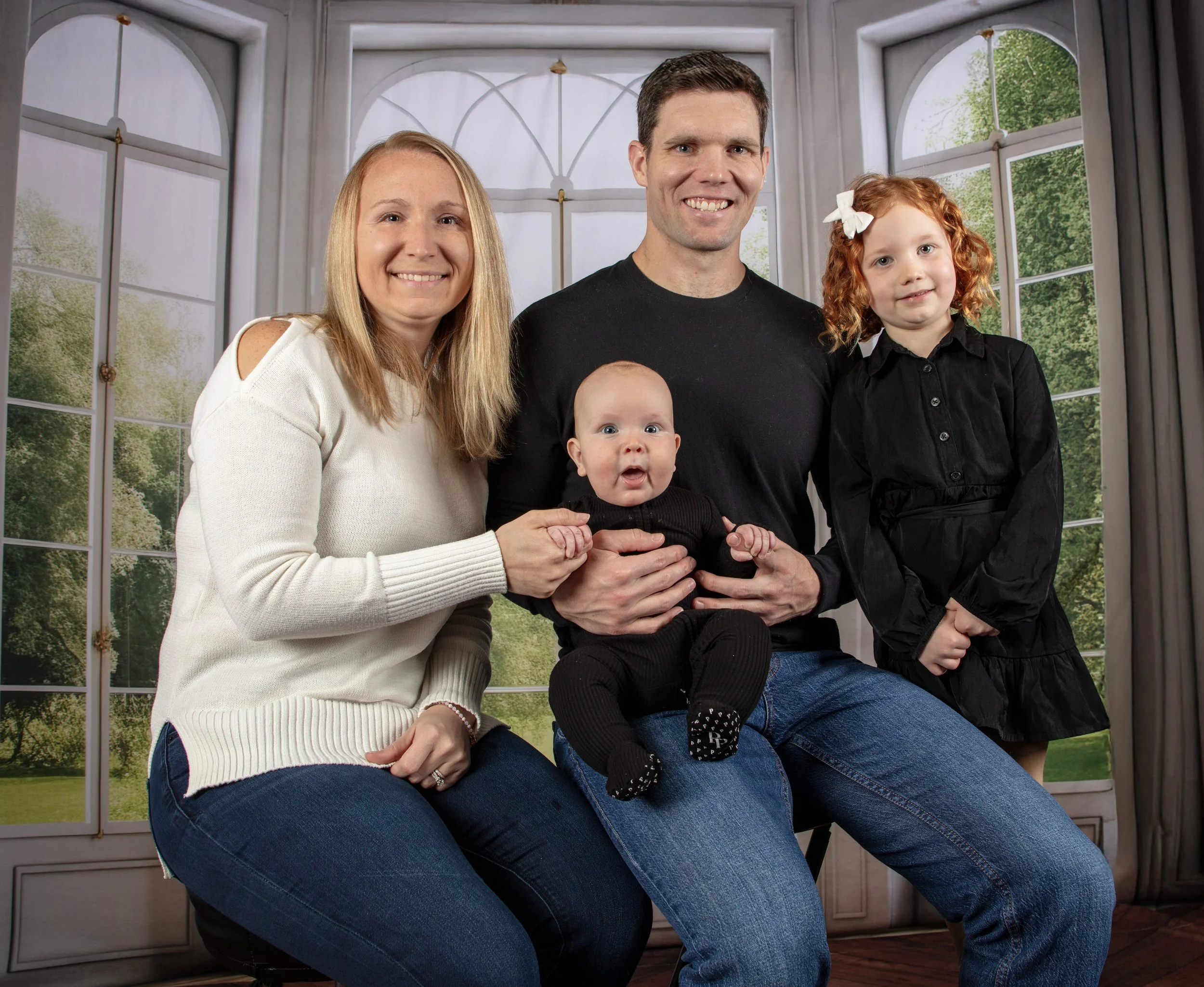 Family portrait of four people, including two women, one man, and two young girls, indoors with large windows showing greenery outside.