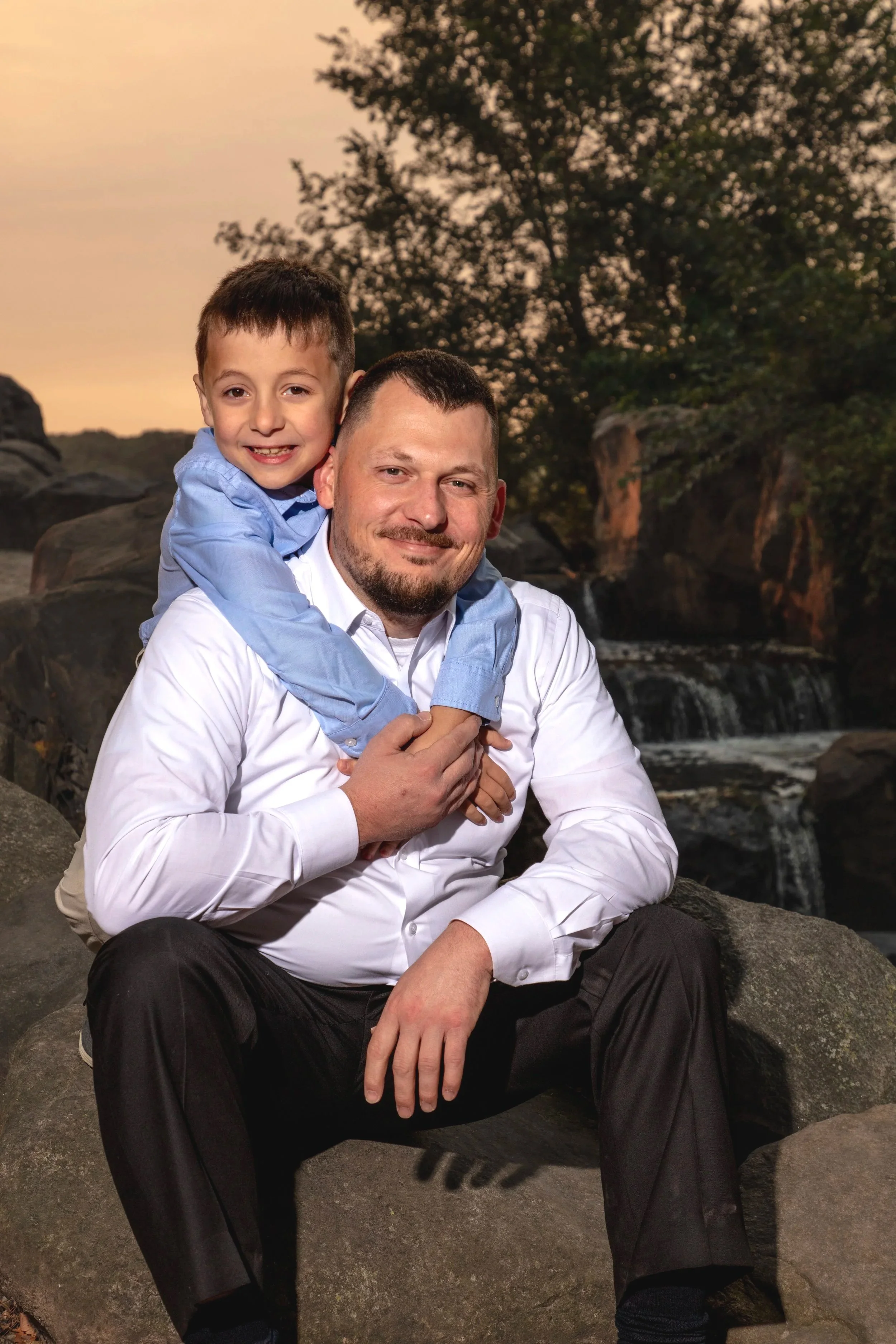 A man and a young boy sitting on rocks outdoors during sunset, with trees and a small waterfall in the background. The boy is hugging the man from behind.