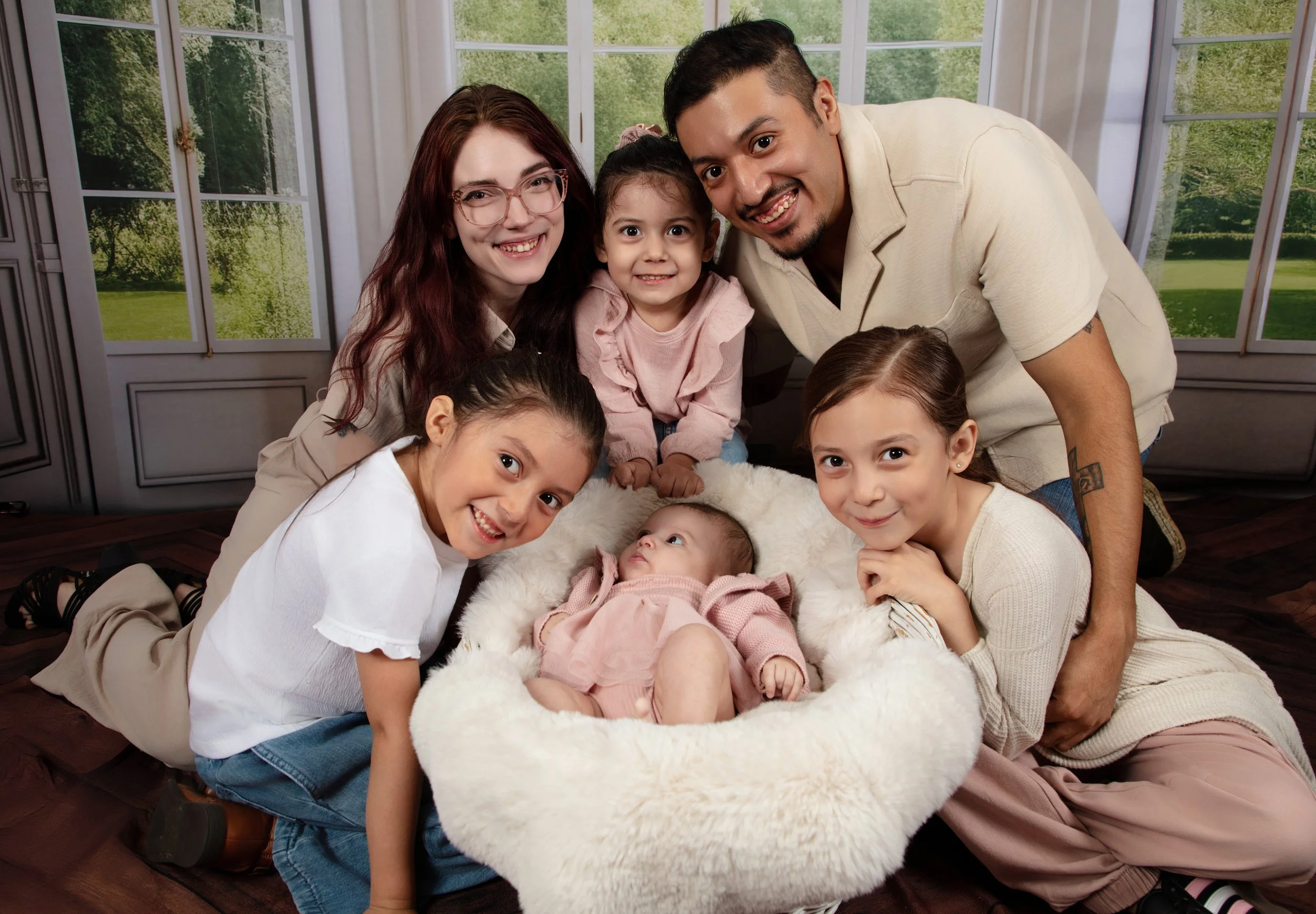 A family of six, including an adult man and woman and four children, gathered around a newborn baby in a cozy room with large windows and green outdoor scenery. They are smiling and posing for the photo.