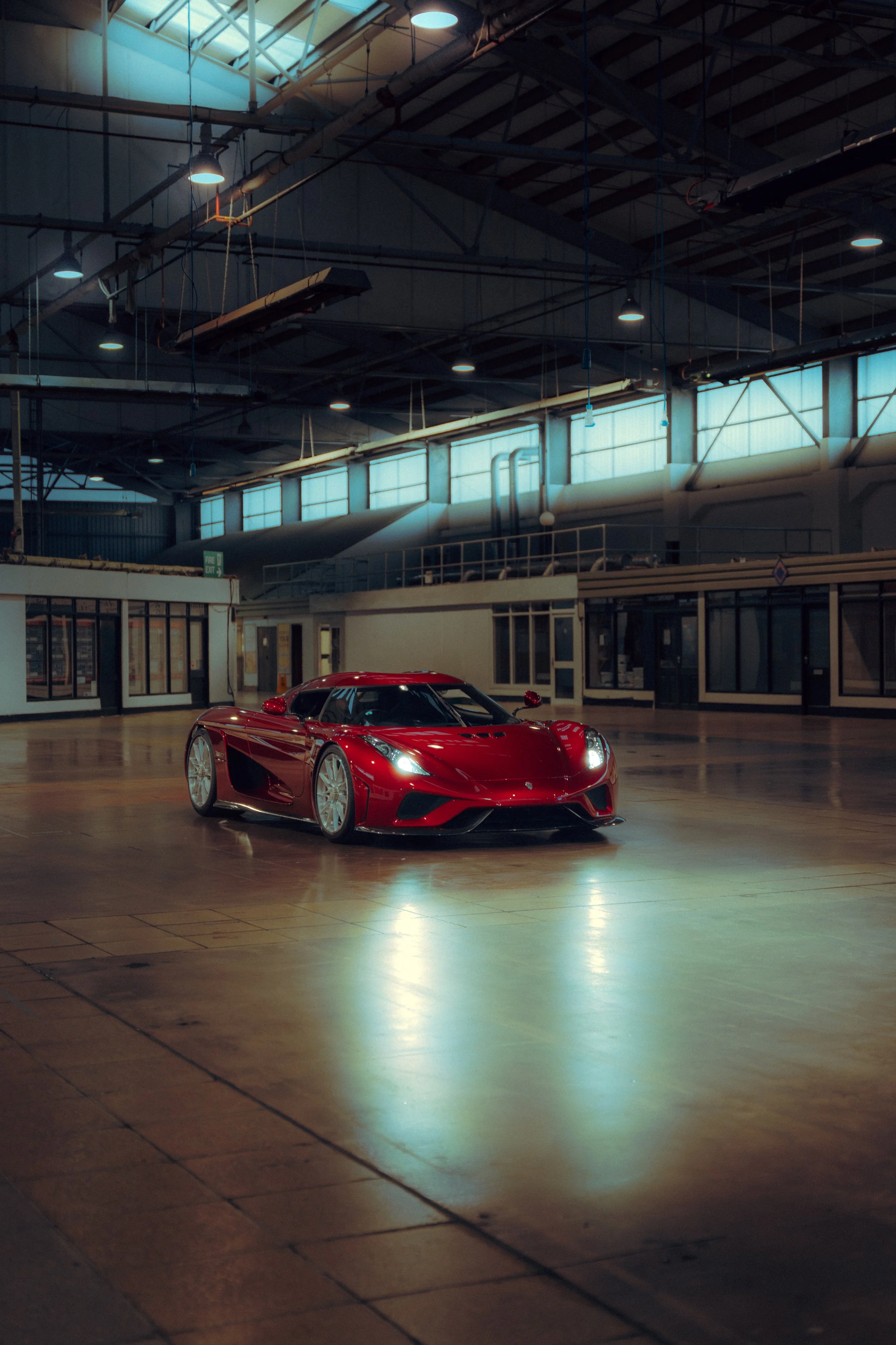 A red Koenigsegg inside a large industrial-style building with high ceilings and skylights, dimly lit with overhead lights.