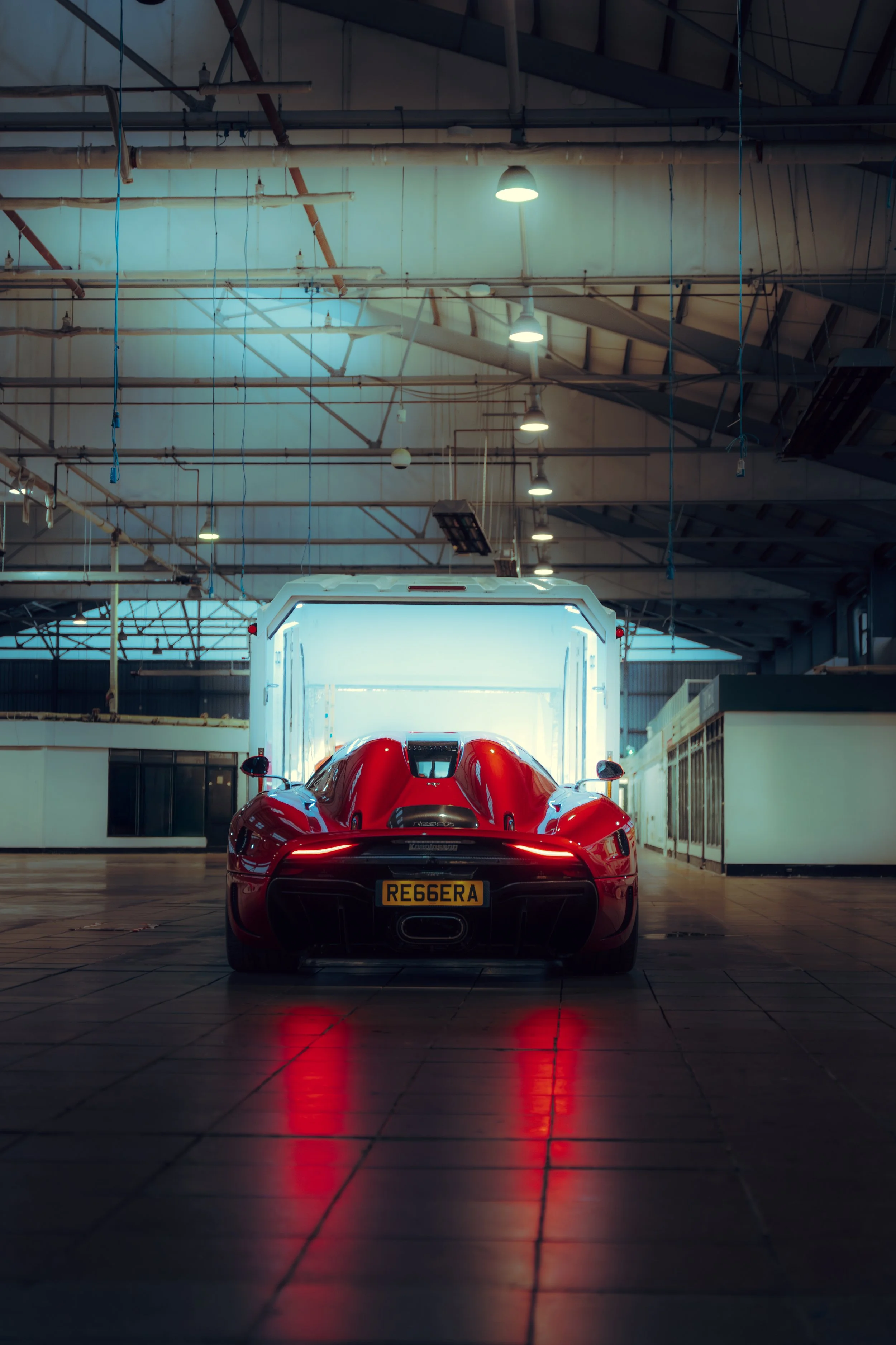 A red sports car is parked inside a garage with bright overhead lighting, license plate number RE66ERA, with reflections on the tiled floor.