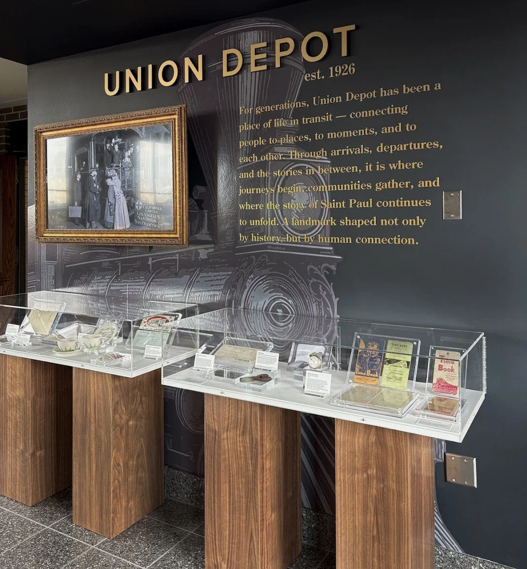 Union Depot display with historical transit items in glass cases; black wall features gold text about the depots history and a vintage photo, with a train illustration in the background.