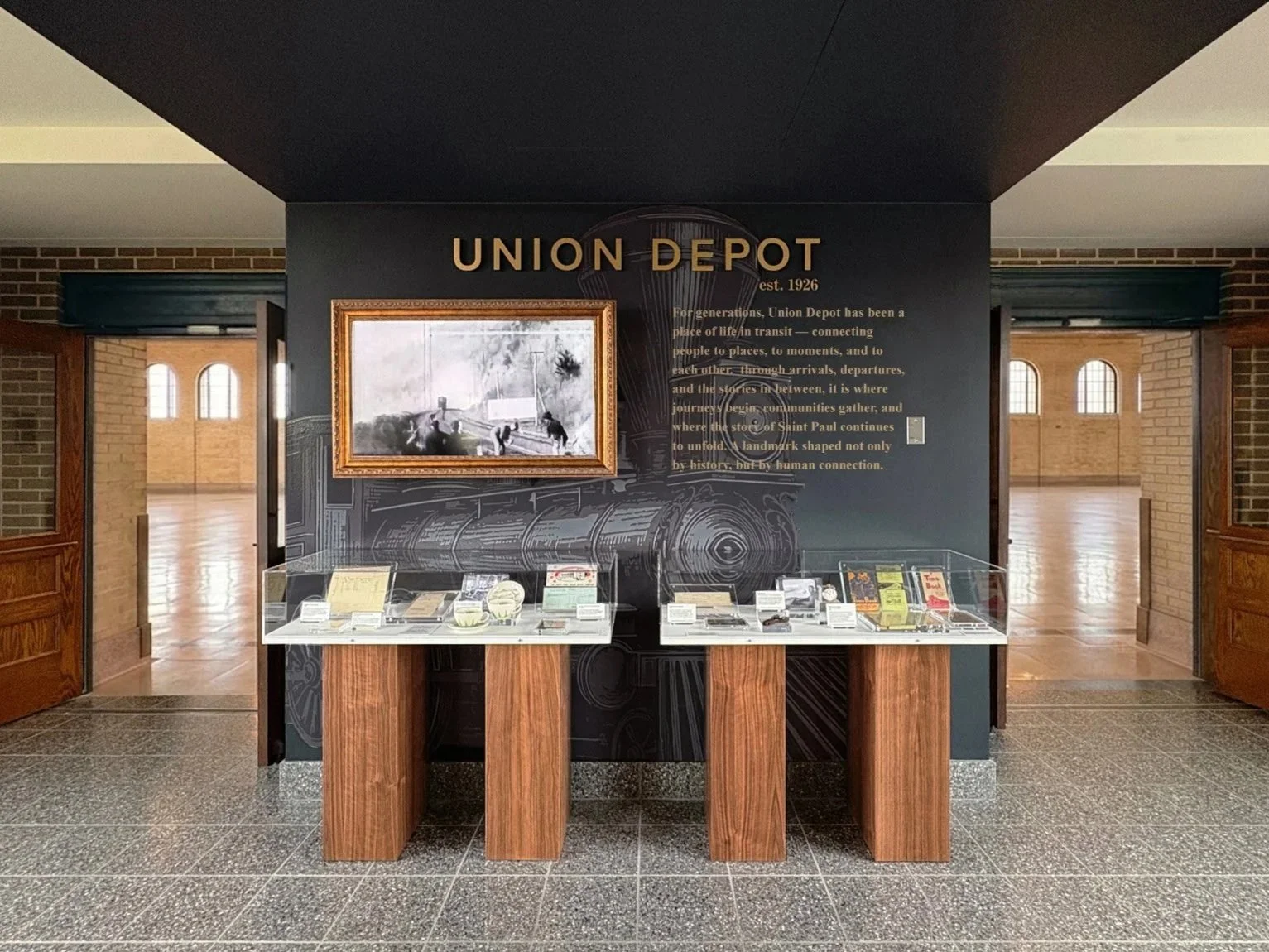 A display at Union Depot features historical artifacts, vintage tickets, and a black-and-white train photo on a wall with informational text. Wooden doors and arched windows frame the bright, spacious room in the background.