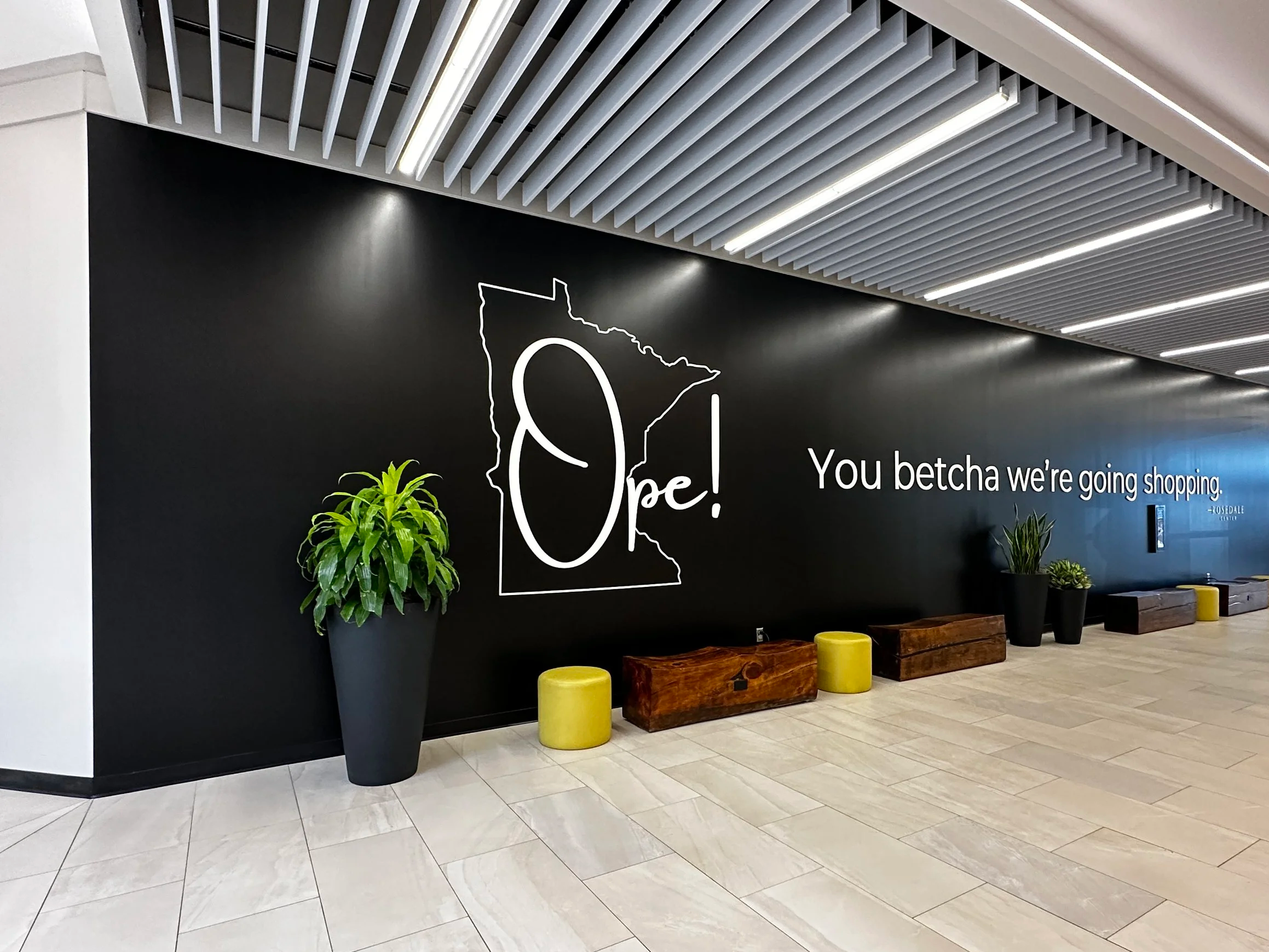 Interior of a modern shopping mall with a black wall featuring white outline of the state of Minnesota and the words "We are Open!" and "You betcha we're going shopping." Several potted plants and wooden benches are arranged along the wall, with polished tiled floors and a ceiling with linear lighting and decorative paneling.