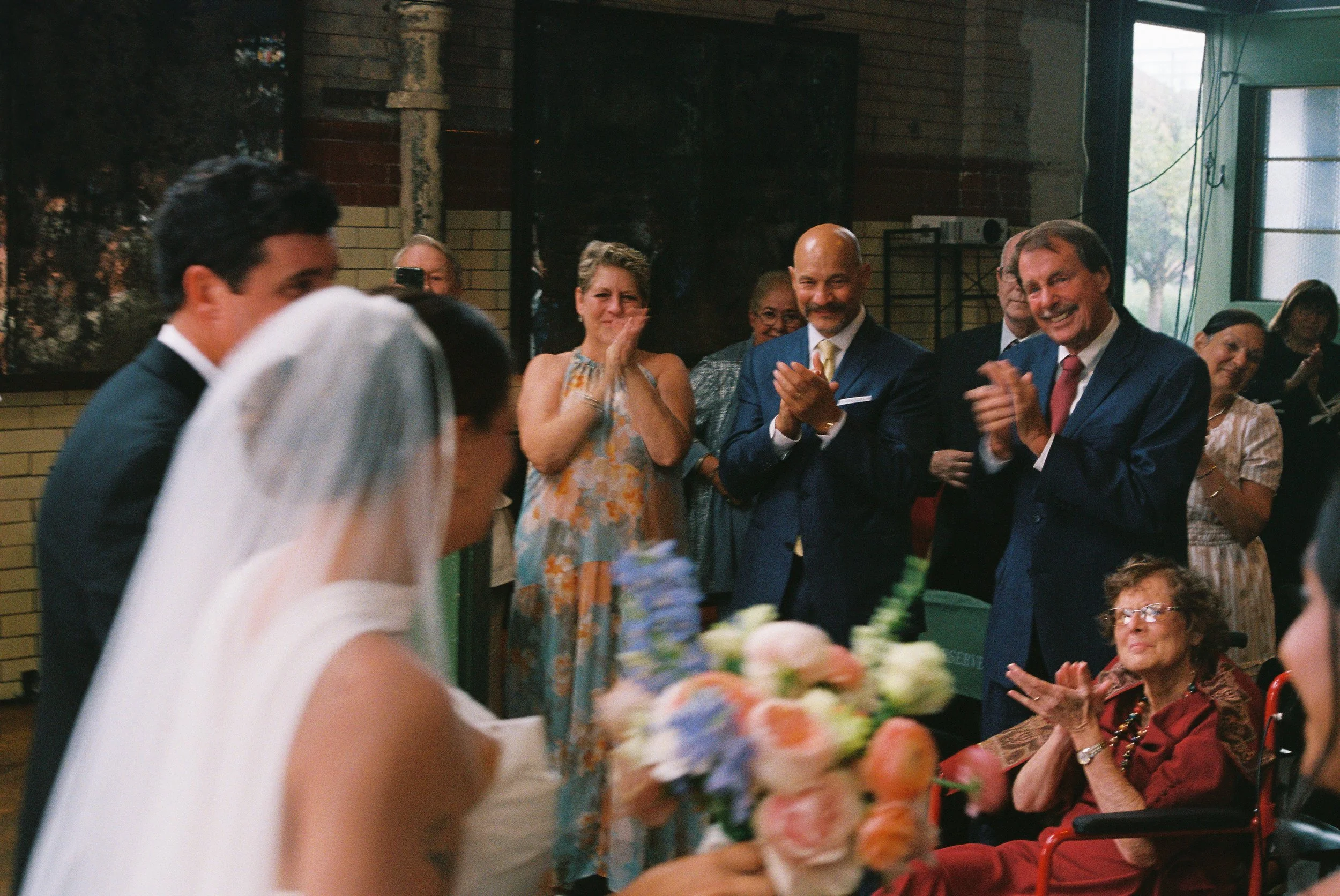 A bride and groom at their wedding ceremony with guests clapping and smiling, including a woman in a red dress in a wheelchair, in an indoor venue with large windows.