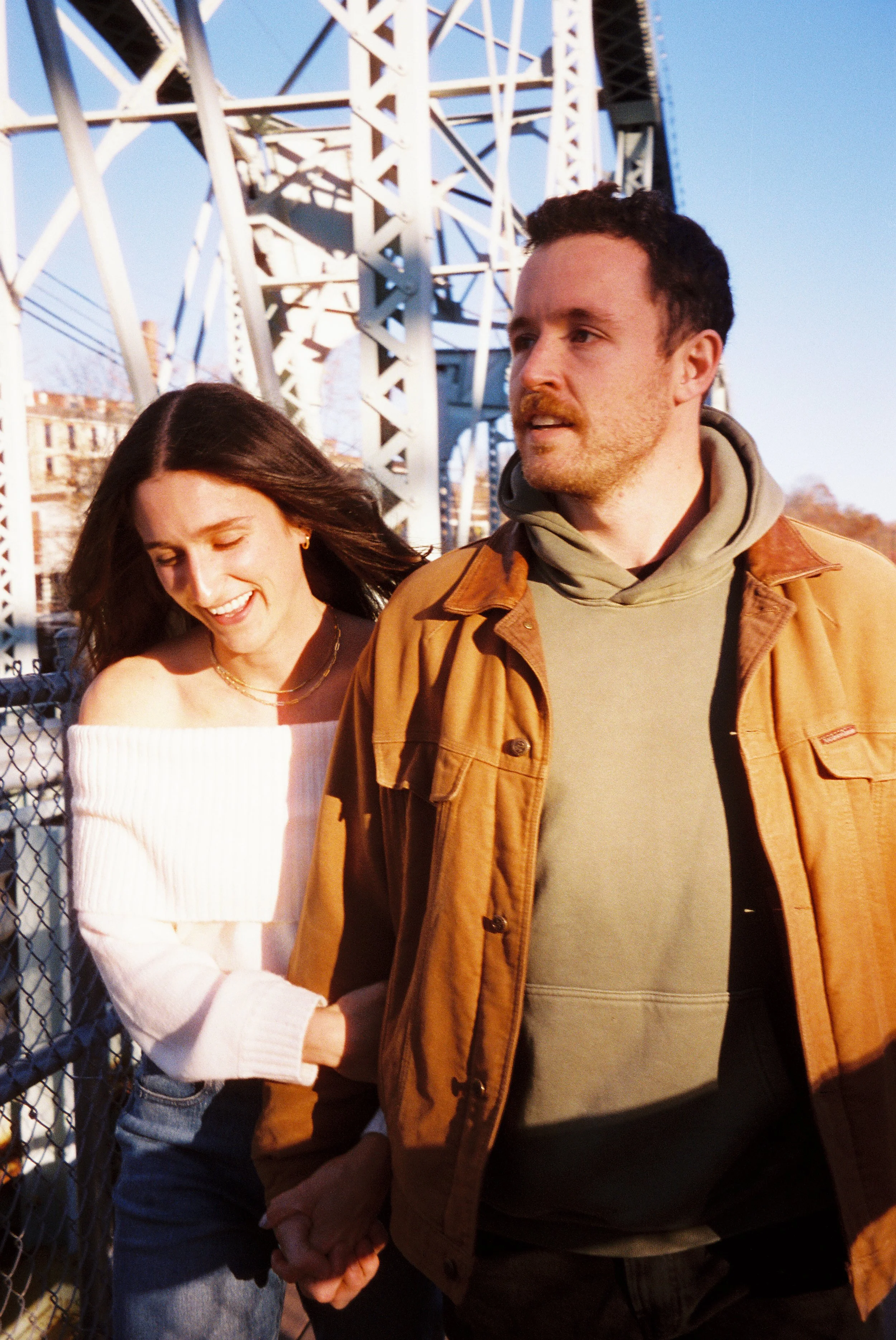 A smiling woman and a man holding hands, walking past a metal bridge structure in the late afternoon sunlight.