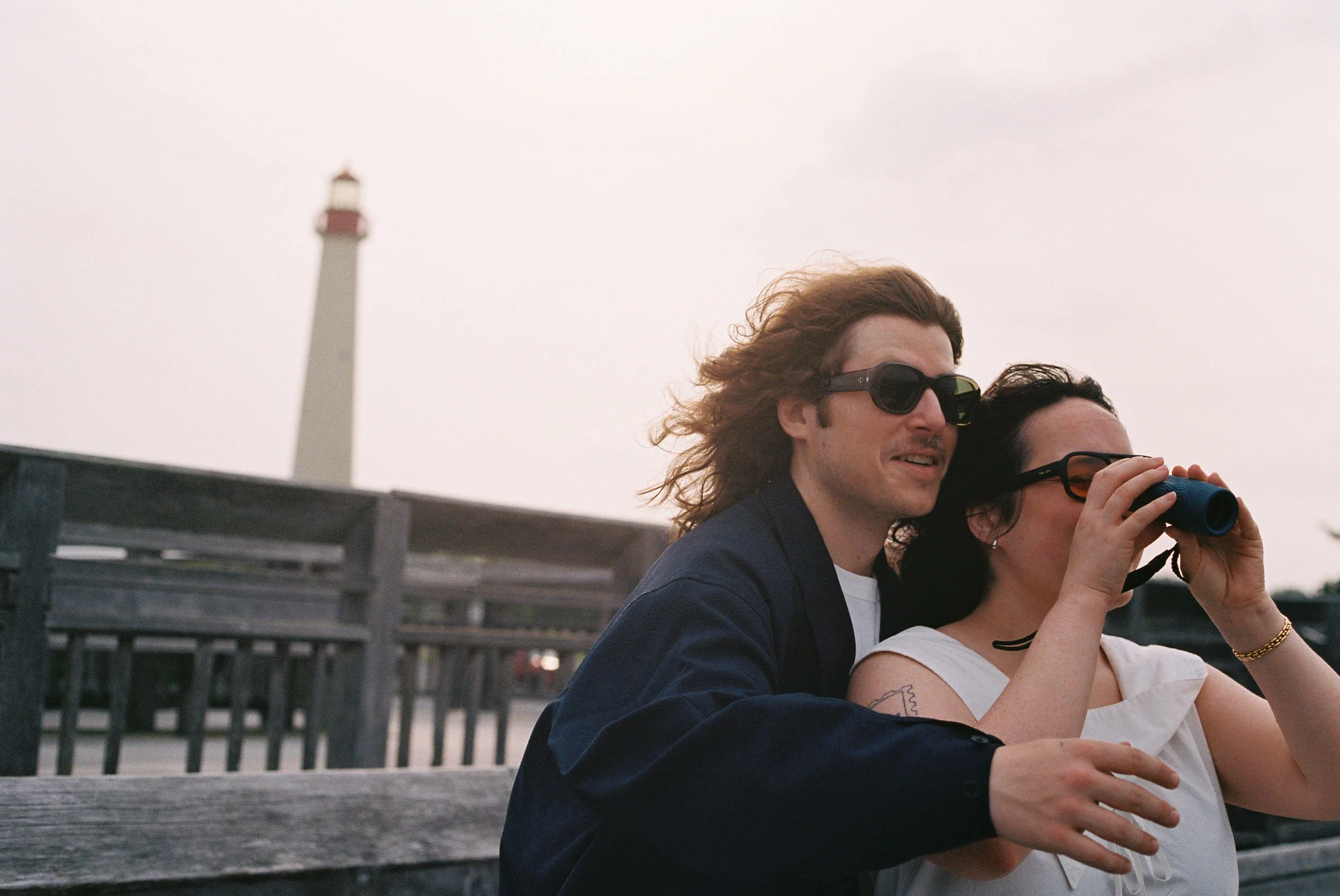 A man and woman sitting outside on a wooden bench, with the man looking into the distance and the woman using binoculars, a lighthouse is in the background.