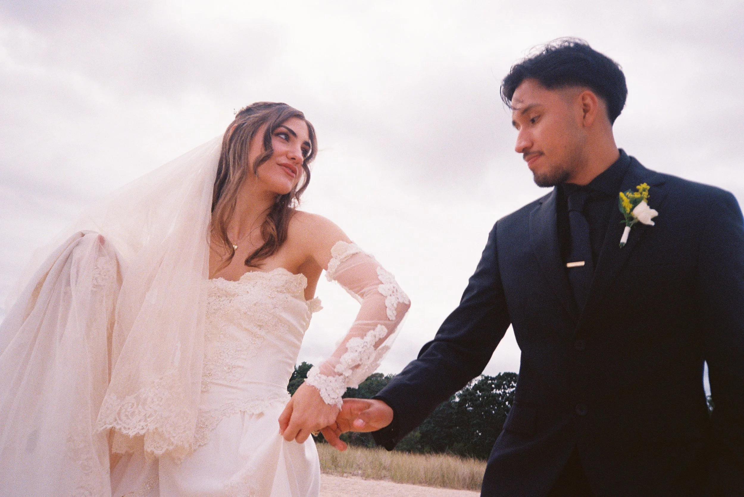 A bride and groom holding hands outdoors on a cloudy day, with the bride in a lace wedding gown and the groom in a dark suit with a boutonnière.