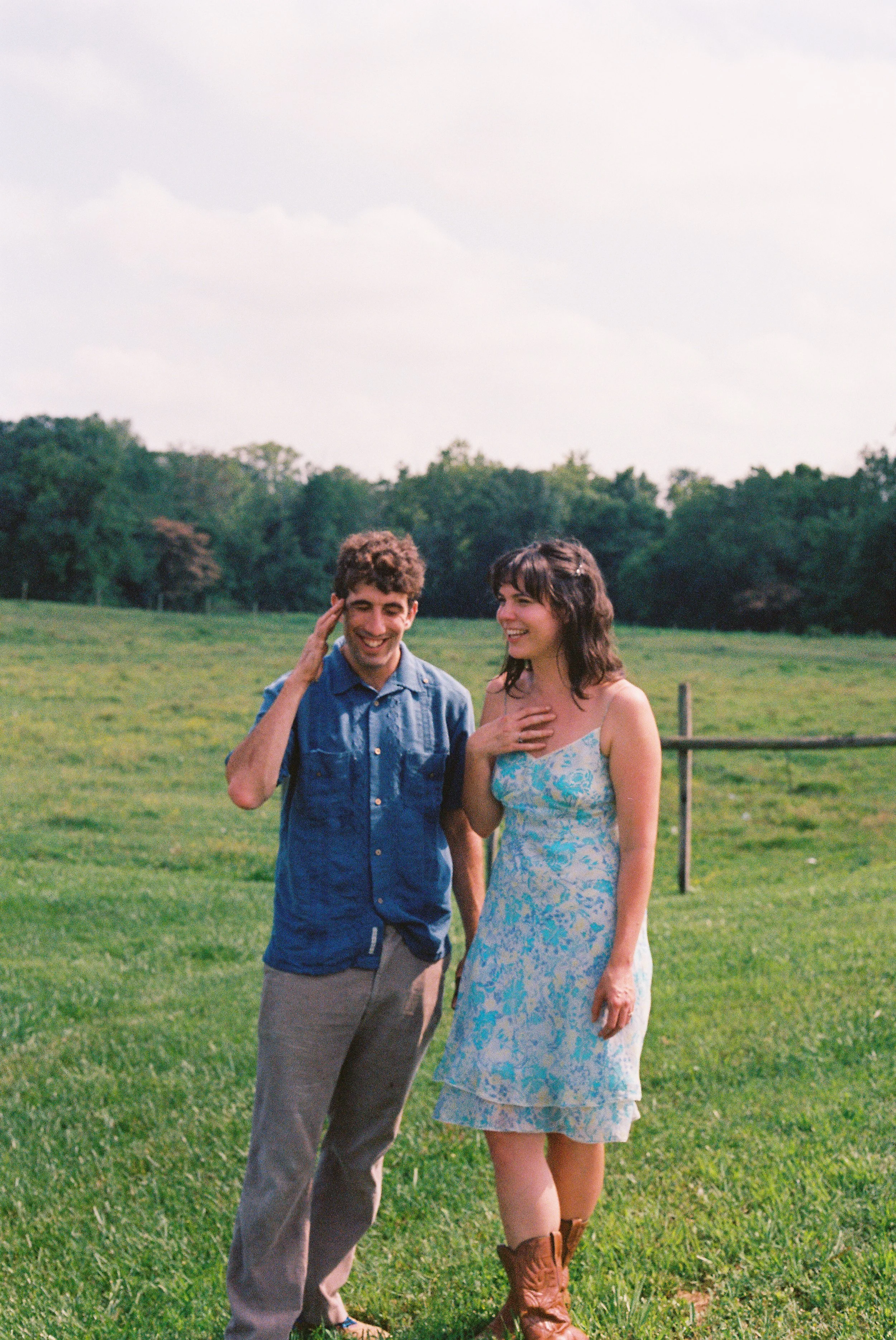 A young man and woman standing in a grassy field, smiling and laughing, with trees and a cloudy sky in the background.