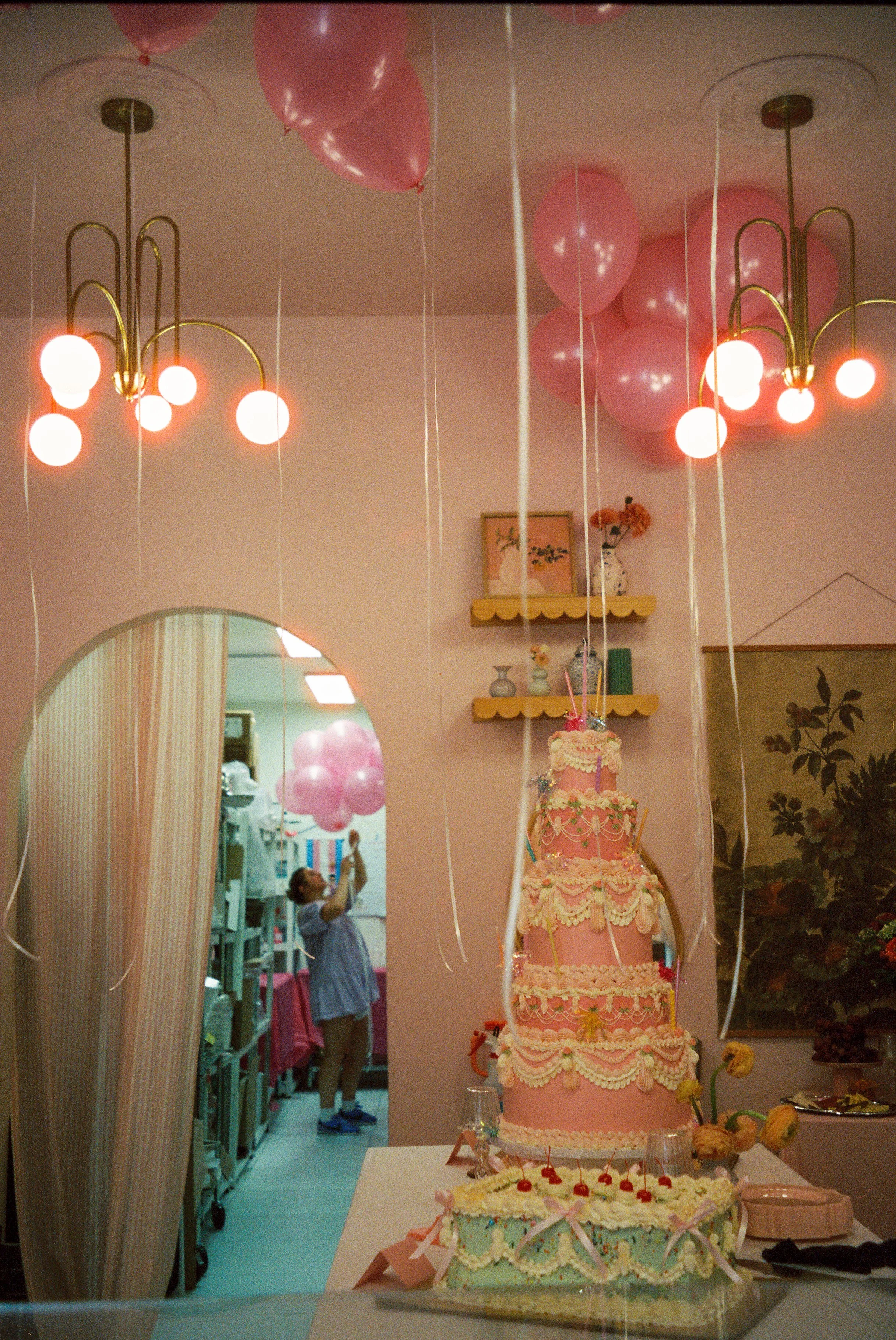 A birthday party scene with a two-tier pink wedding cake decorated with cream-colored icing and flowers, a smaller cake with cherries on top in front of it, pink balloons on the ceiling, and a young child in the background holding pink balloons.