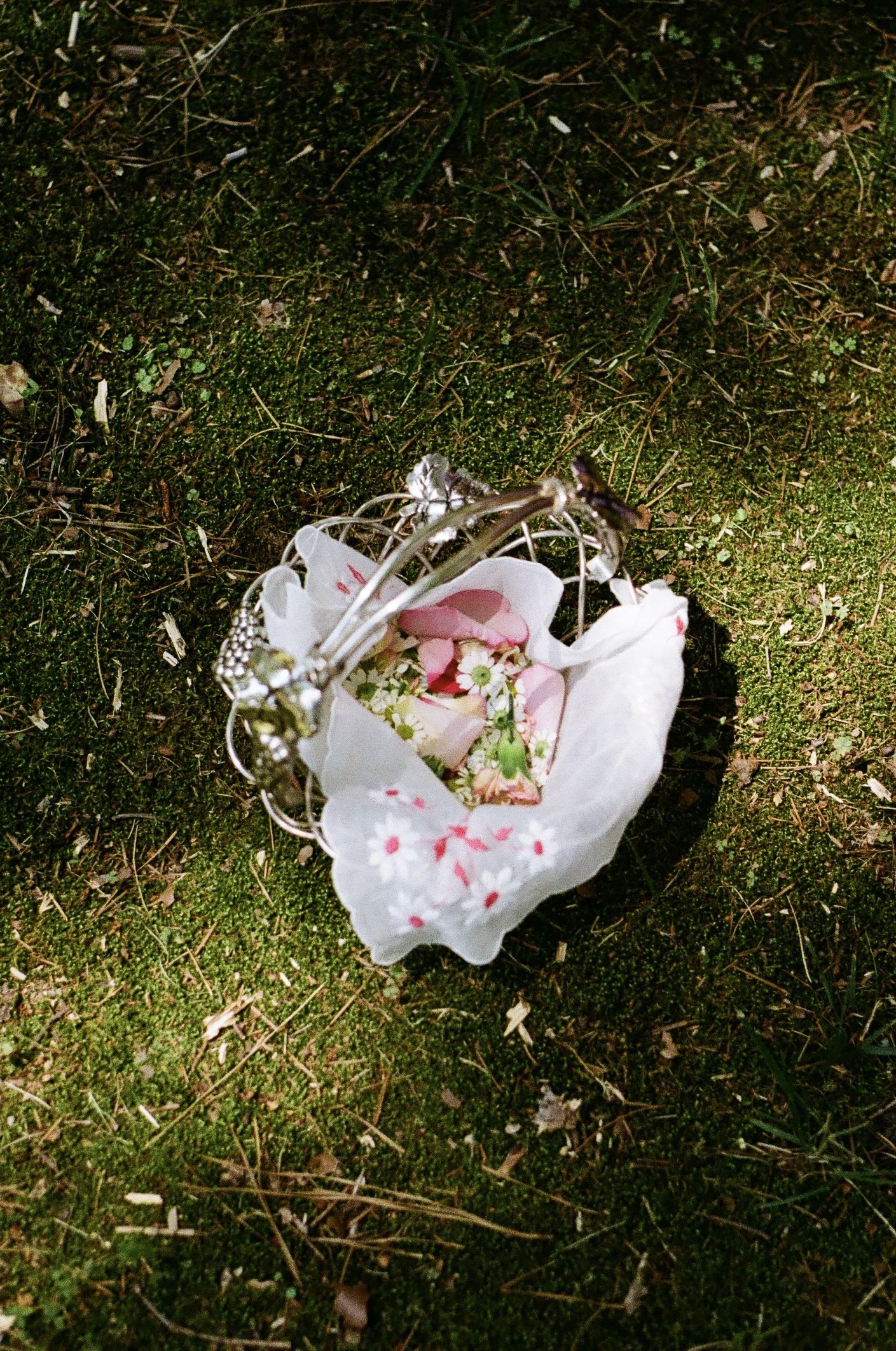 A small bouquet of pink and white flowers wrapped in white paper, resting on mossy ground.