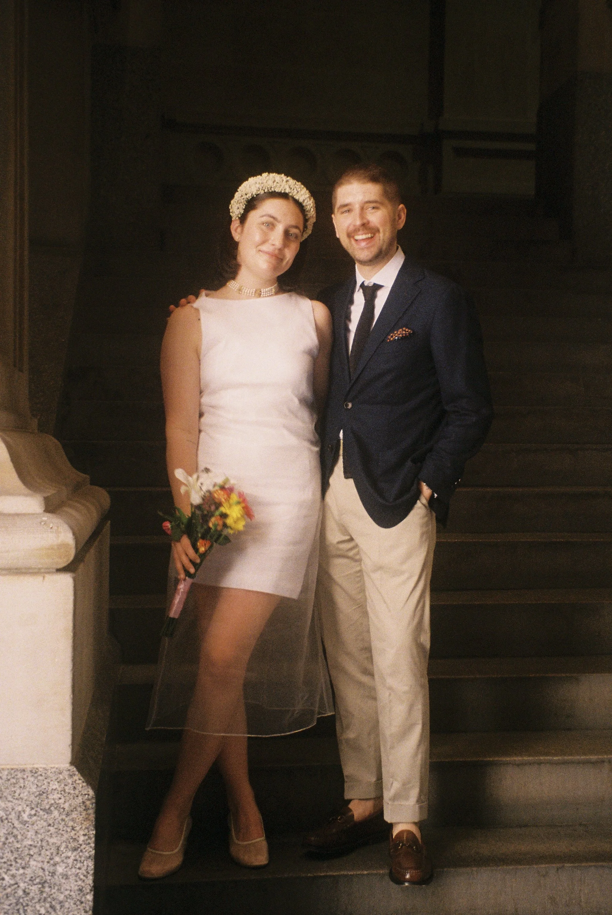 A woman in a white dress with a pearl headband and veil holding a bouquet, standing beside a man in a navy blazer, white shirt, and beige pants, on a staircase.