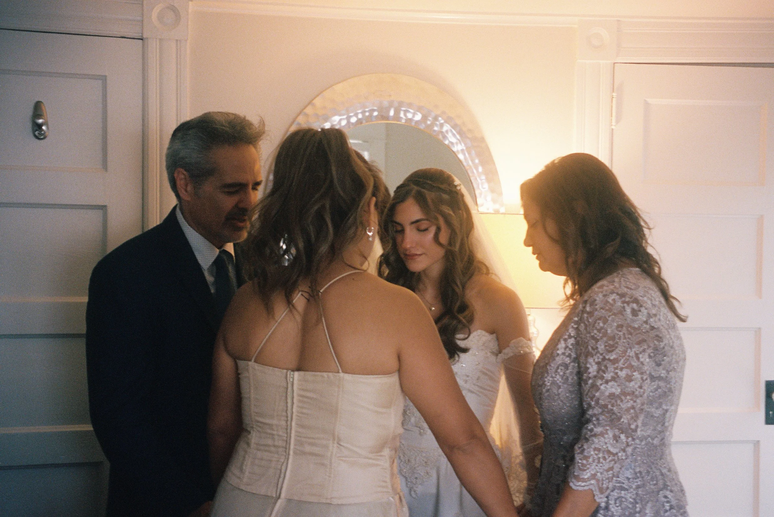 Four people, likely a family, praying together with bowed heads in a solemn moment, possibly at a wedding. They are standing indoors in a warmly lit room with white walls and a mirror in the background.