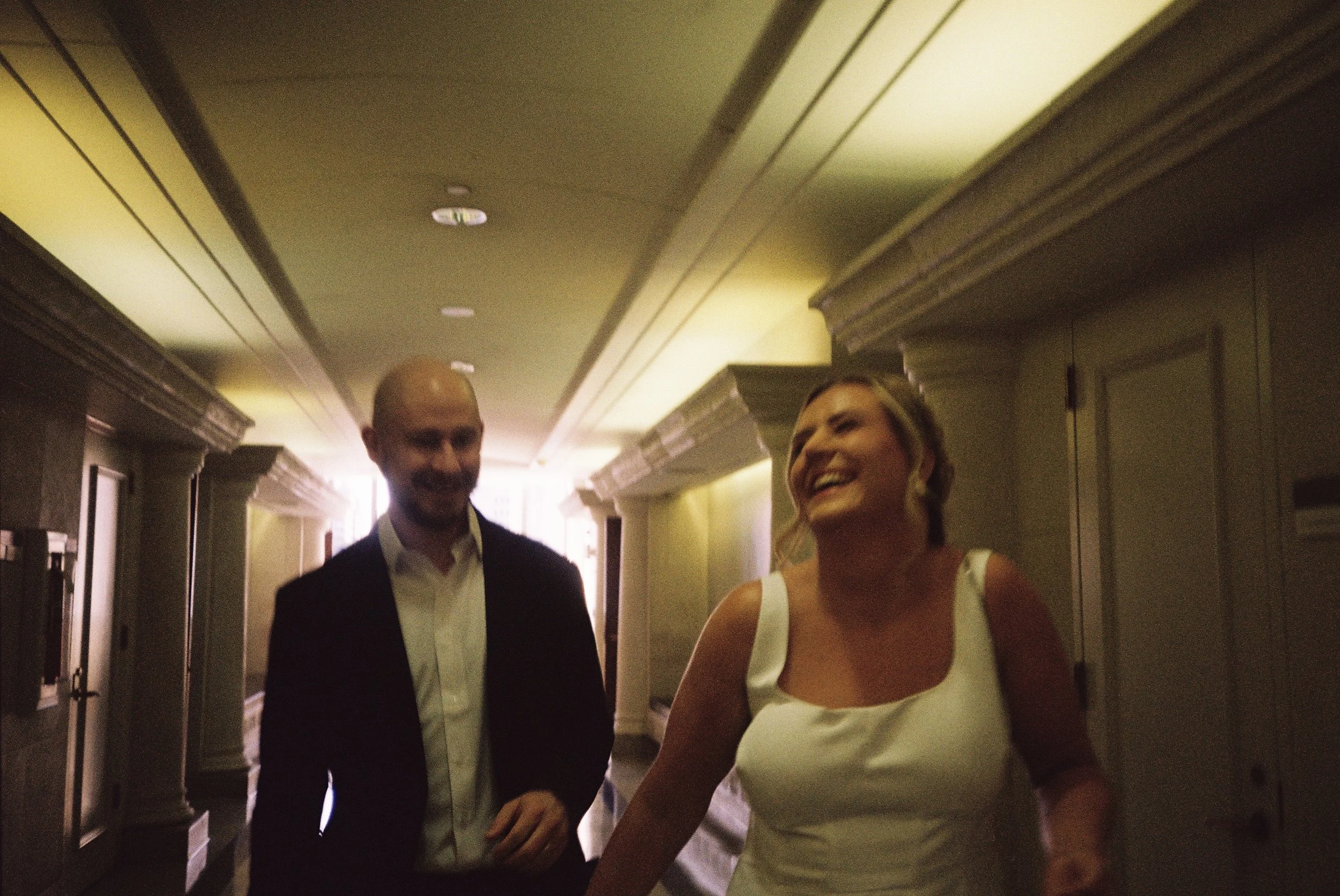 Two people, a man in a tuxedo and a woman in a white dress, are walking down a hallway, smiling and laughing.