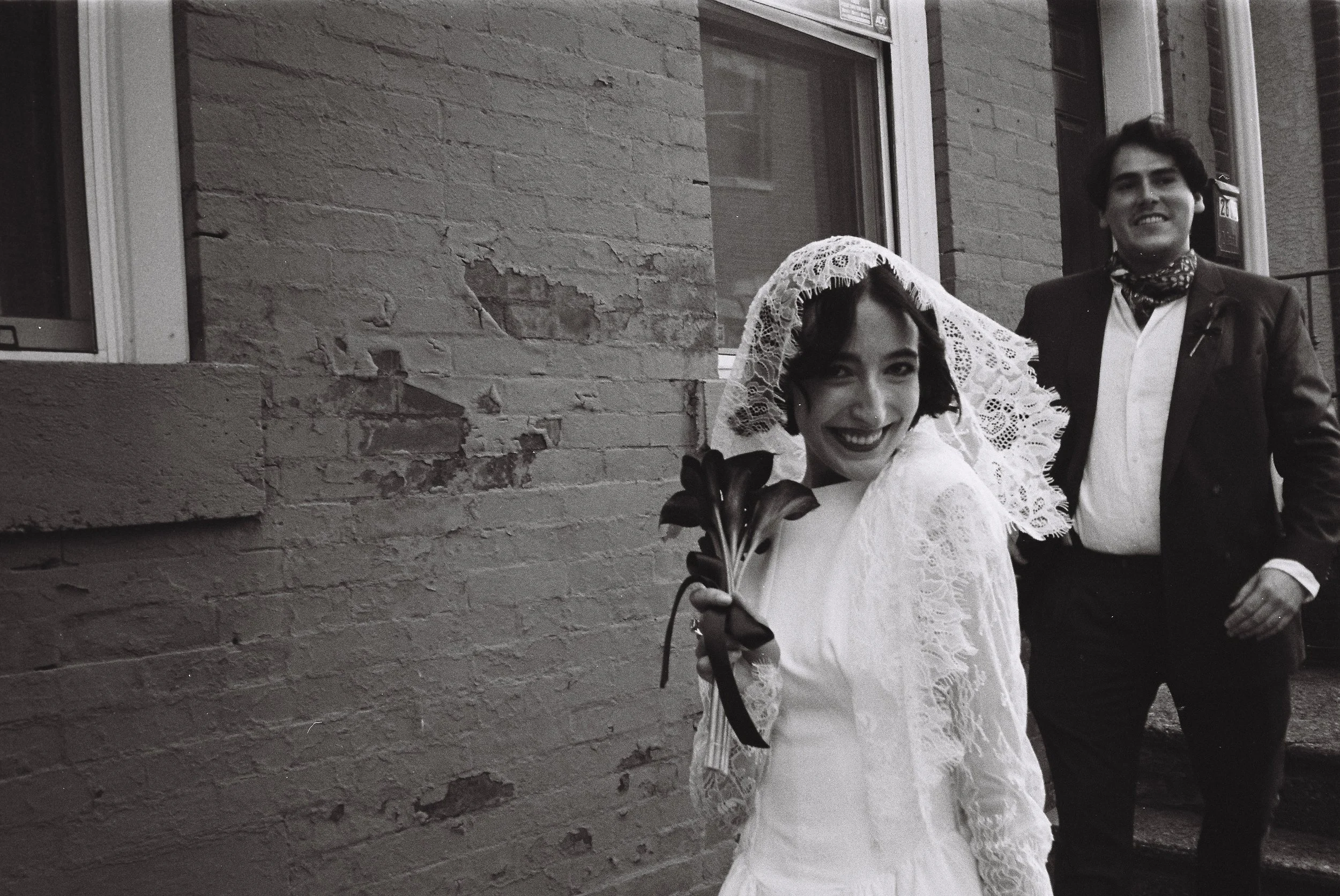 A woman in a wedding dress and lace veil smiling holding a bouquet of flowers, standing outside a brick building, with a man in a suit behind her.
