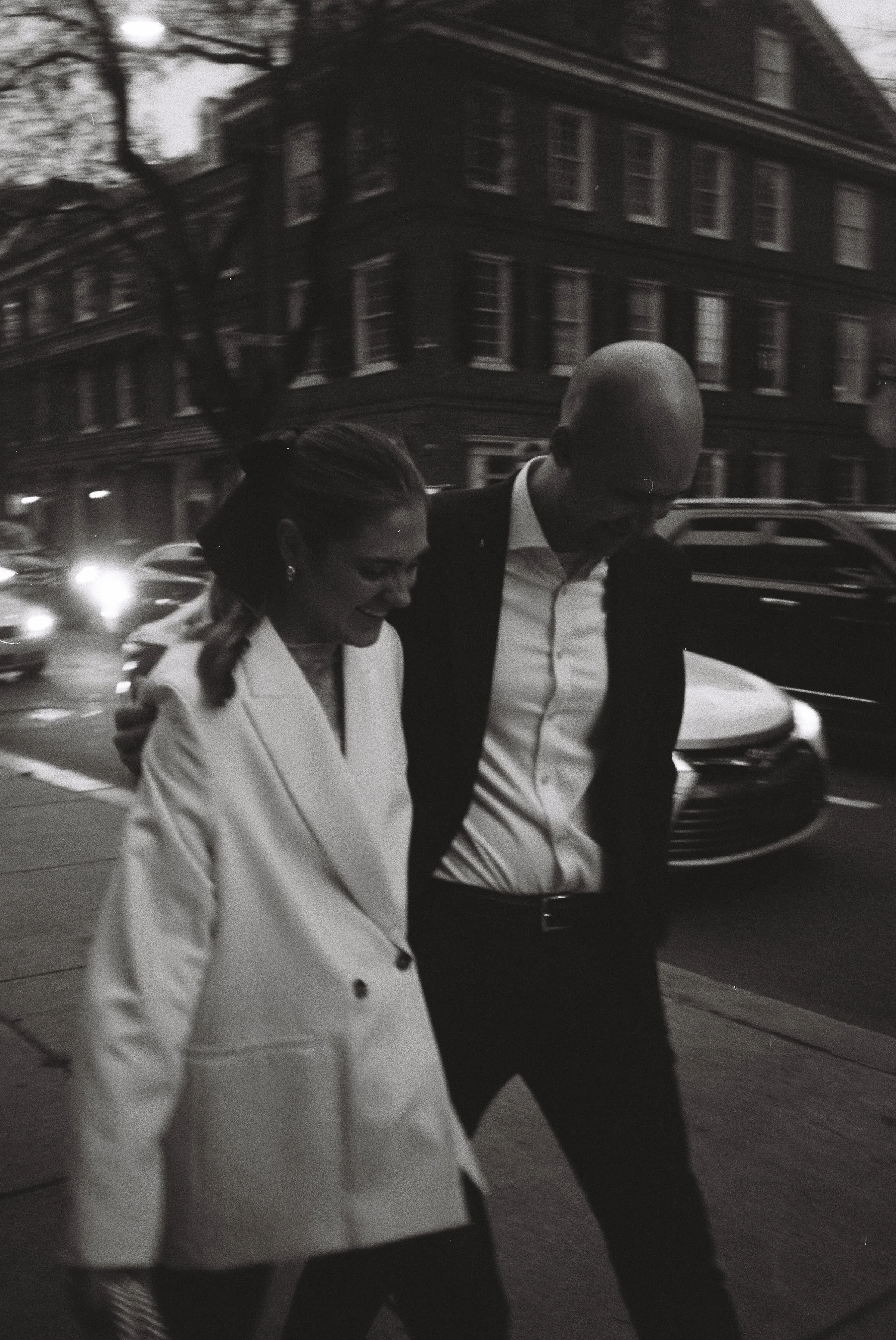 A black-and-white photo of two people walking close together on a city sidewalk, with cars and buildings in the background.