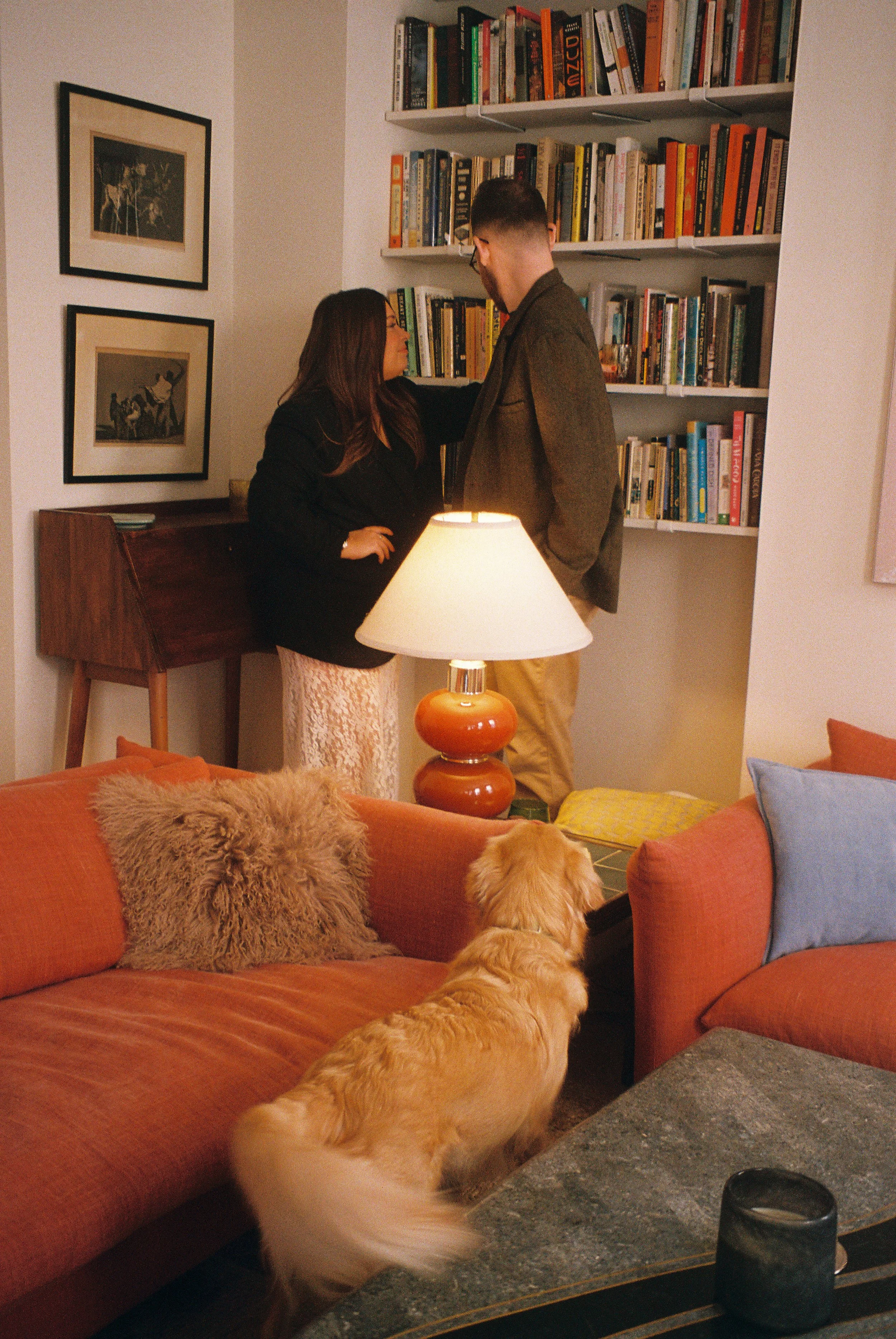 A couple standing close to each other near a bookshelf in a cozy living room, with a dog sitting on the sofa watching them. There is a lamp on a side table in the foreground.
