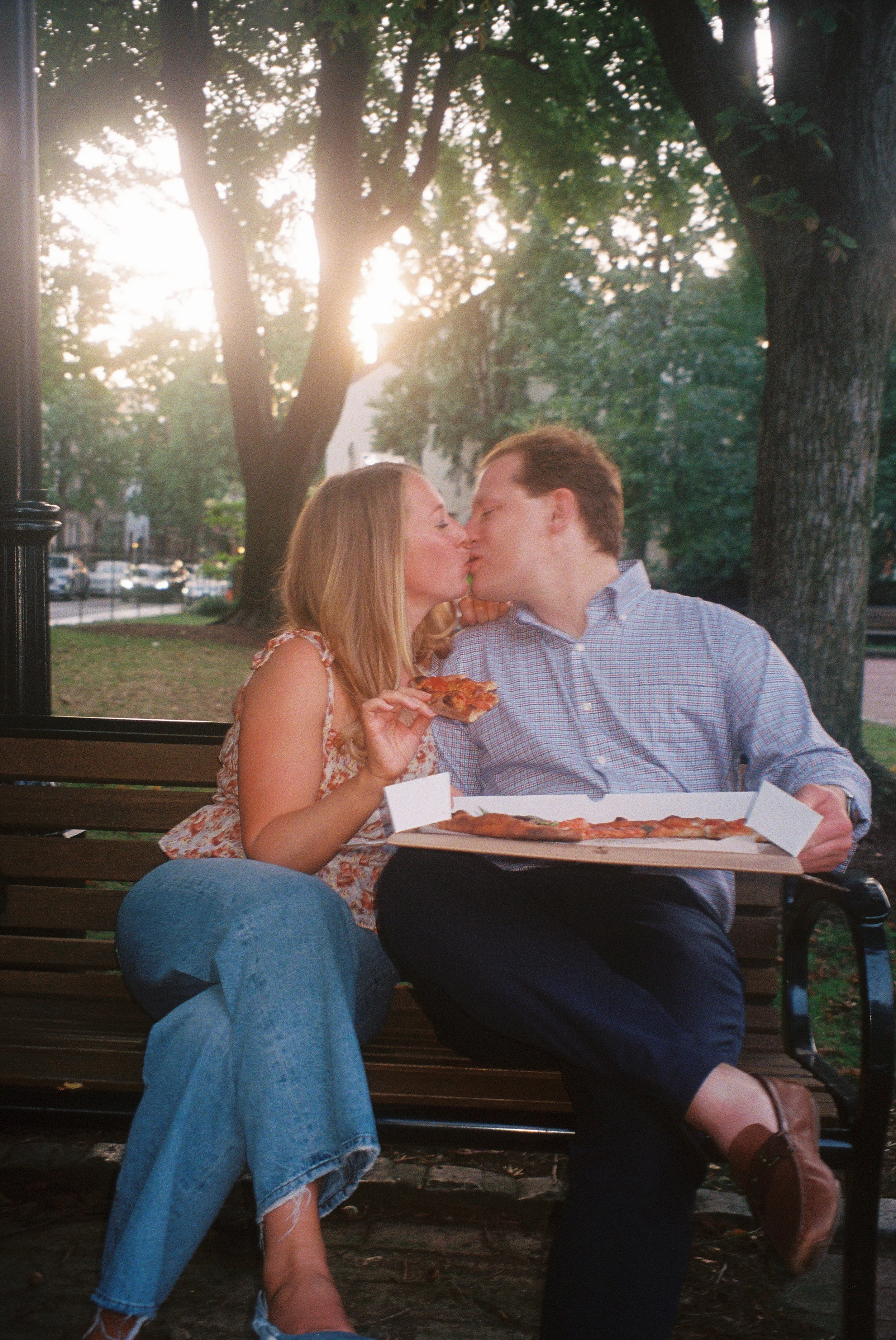 A couple sitting on a park bench sharing a kiss, with pizza in front of them. The woman is holding a slice of pizza, and the man is holding a pizza box. The scene is set outdoors with trees and sunlight in the background.
