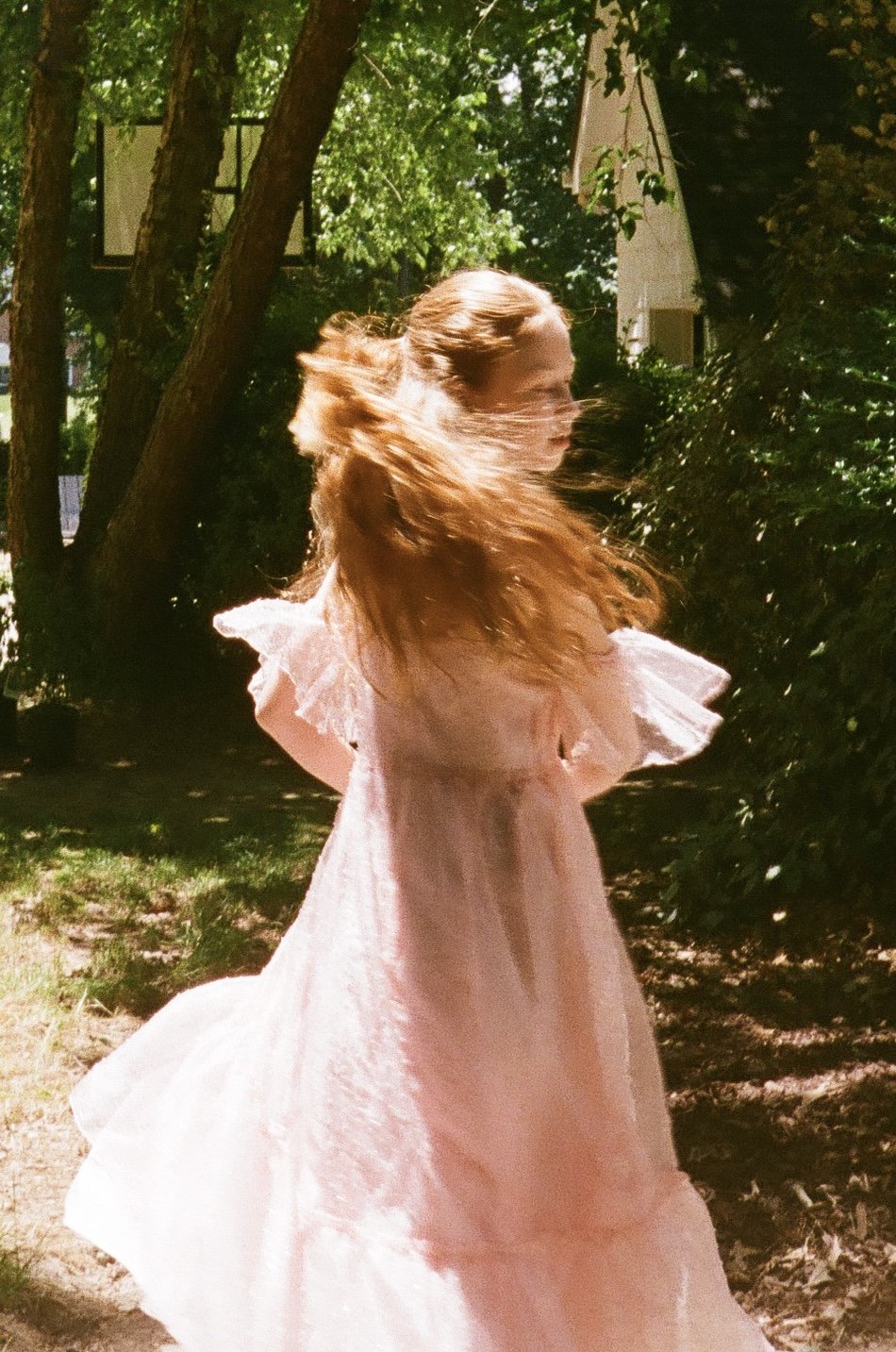 A woman in a light pink dress standing outdoors with trees and sunlight around her, her long hair flowing as she turns her head.
