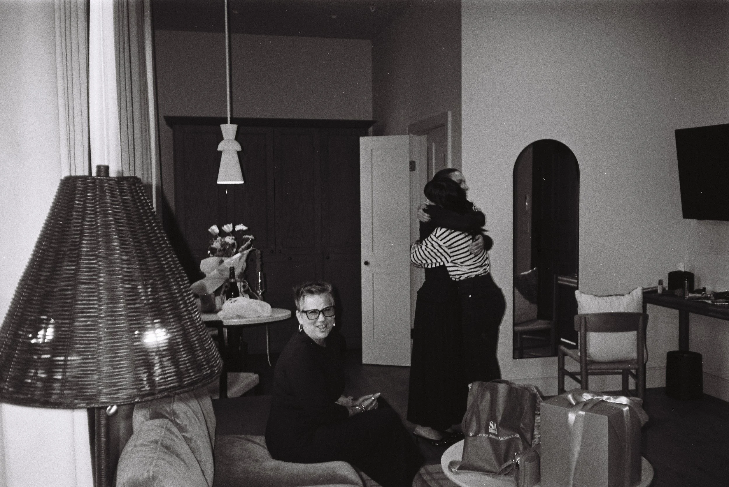Three women in a living room, two are embracing and one is sitting on a couch smiling at the camera. The room has a table with bottles and bags, a lamp, a wall mirror, and a TV.