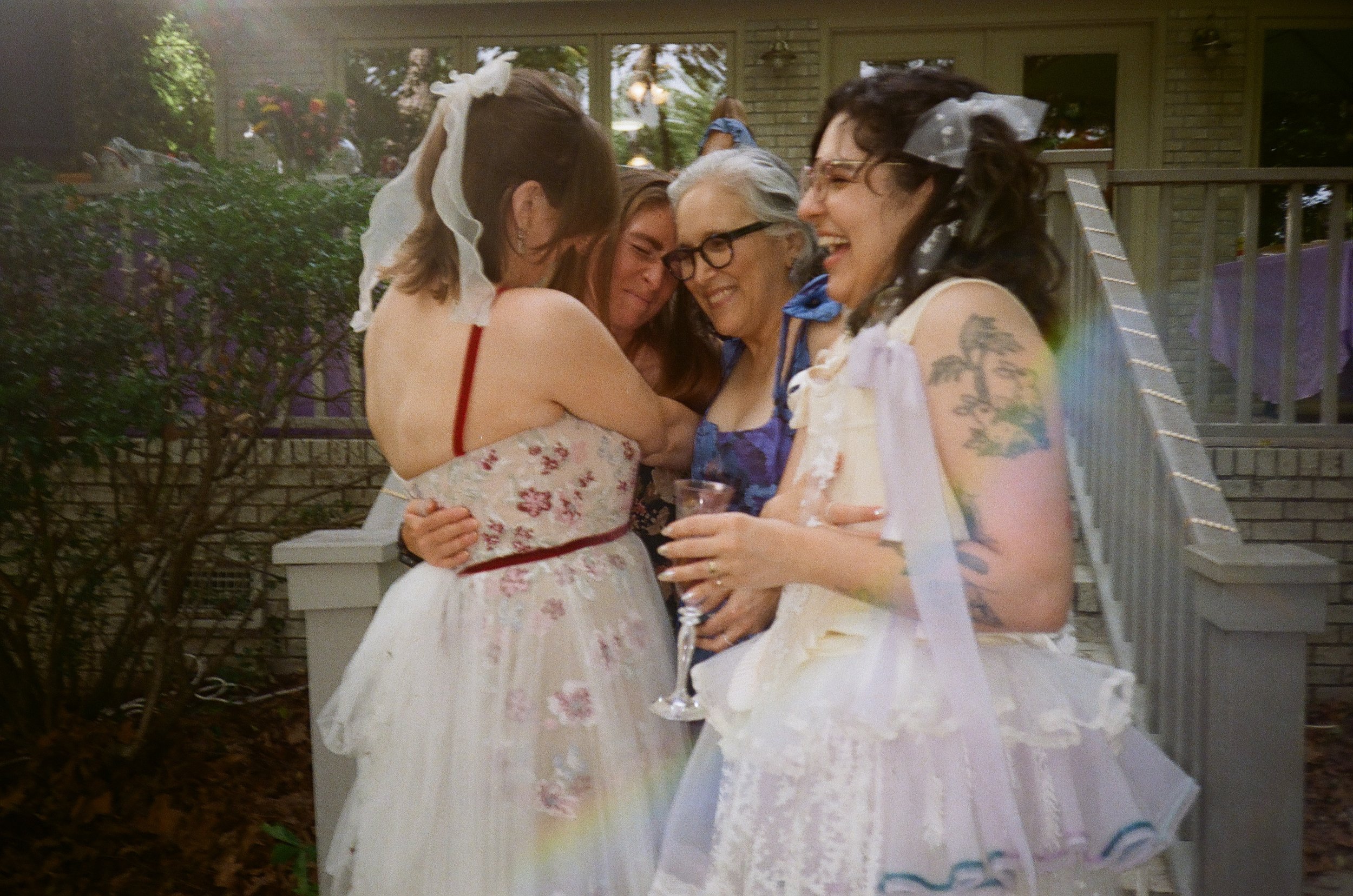 Four women, dressed in vintage or handmade dresses, affectionately hug and smile at a gathering outside on a porch or patio, surrounded by greenery and a house in the background.