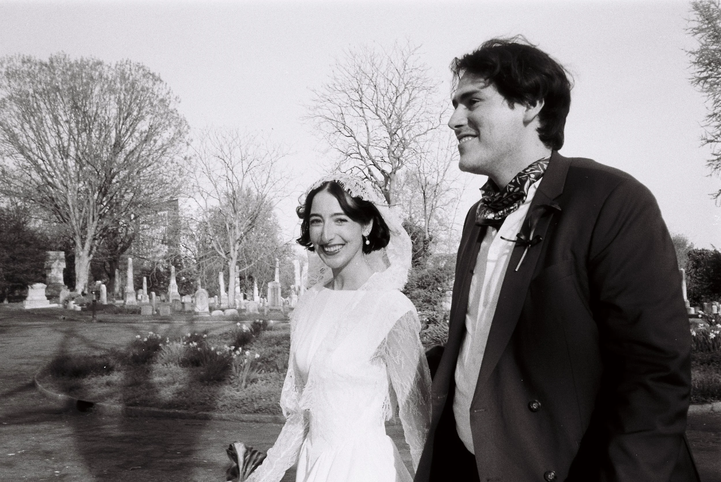 A black and white photo of a bride and groom walking through a cemetery, smiling and looking happy.