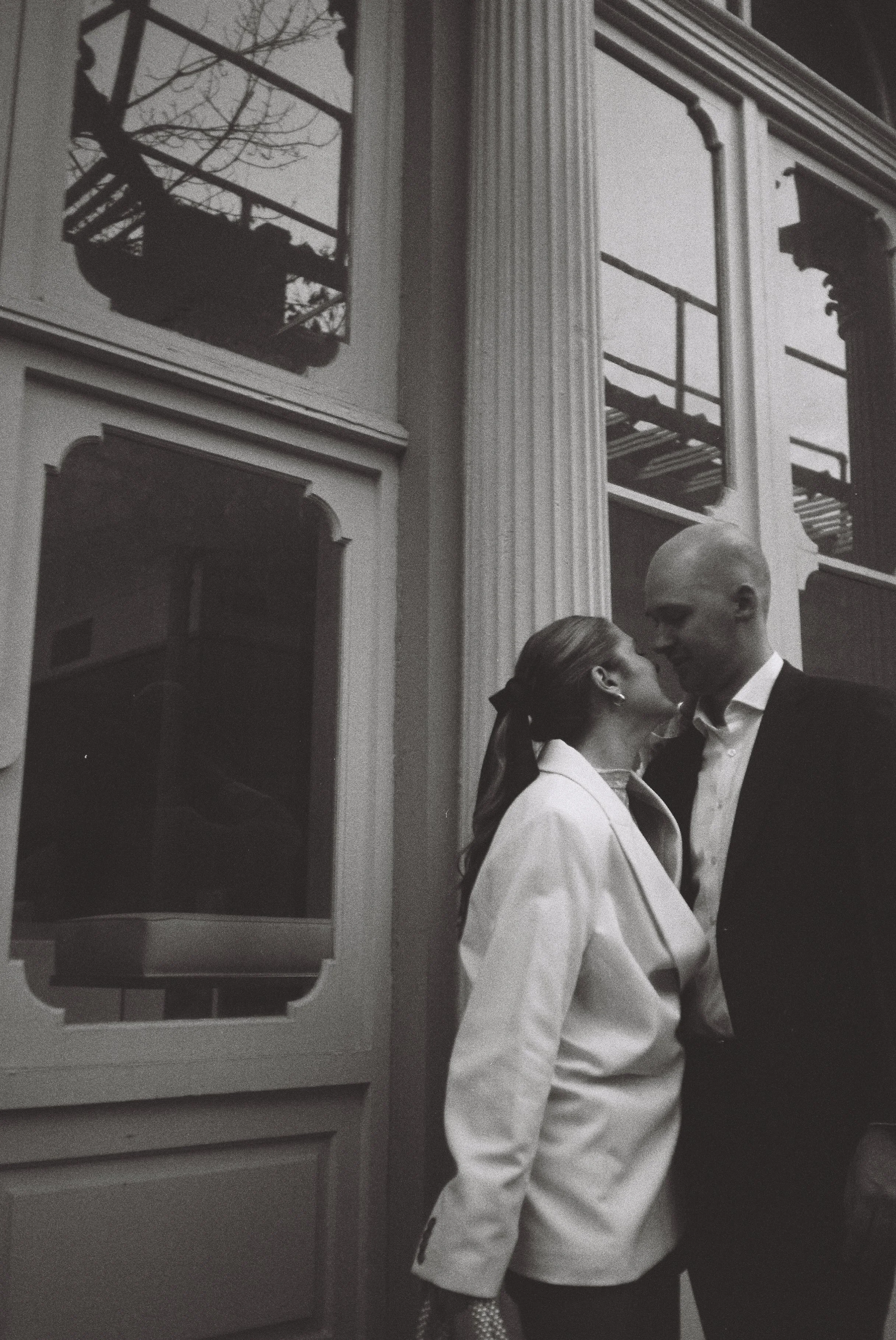 A black and white photo of a man and woman standing close, about to kiss, near a large window with multilevel balconies. The woman is wearing a white blazer and has her hair tied back, while the man is in a dark suit.