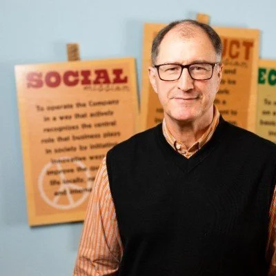 Headshot of Rob Michalak wearing an orange and white stripe button down, a black sweater vest, and black framed glasses. There are protest signs in the background with blurry writing and a visible peace sign.