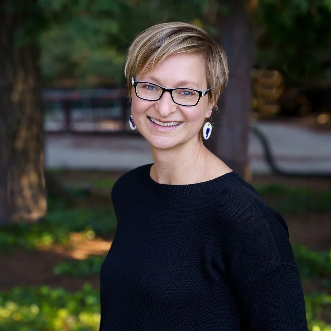 Headshot of Shauna Sadowski wearing a black long sleeve shirt, black framed glasses, and shell earrings.