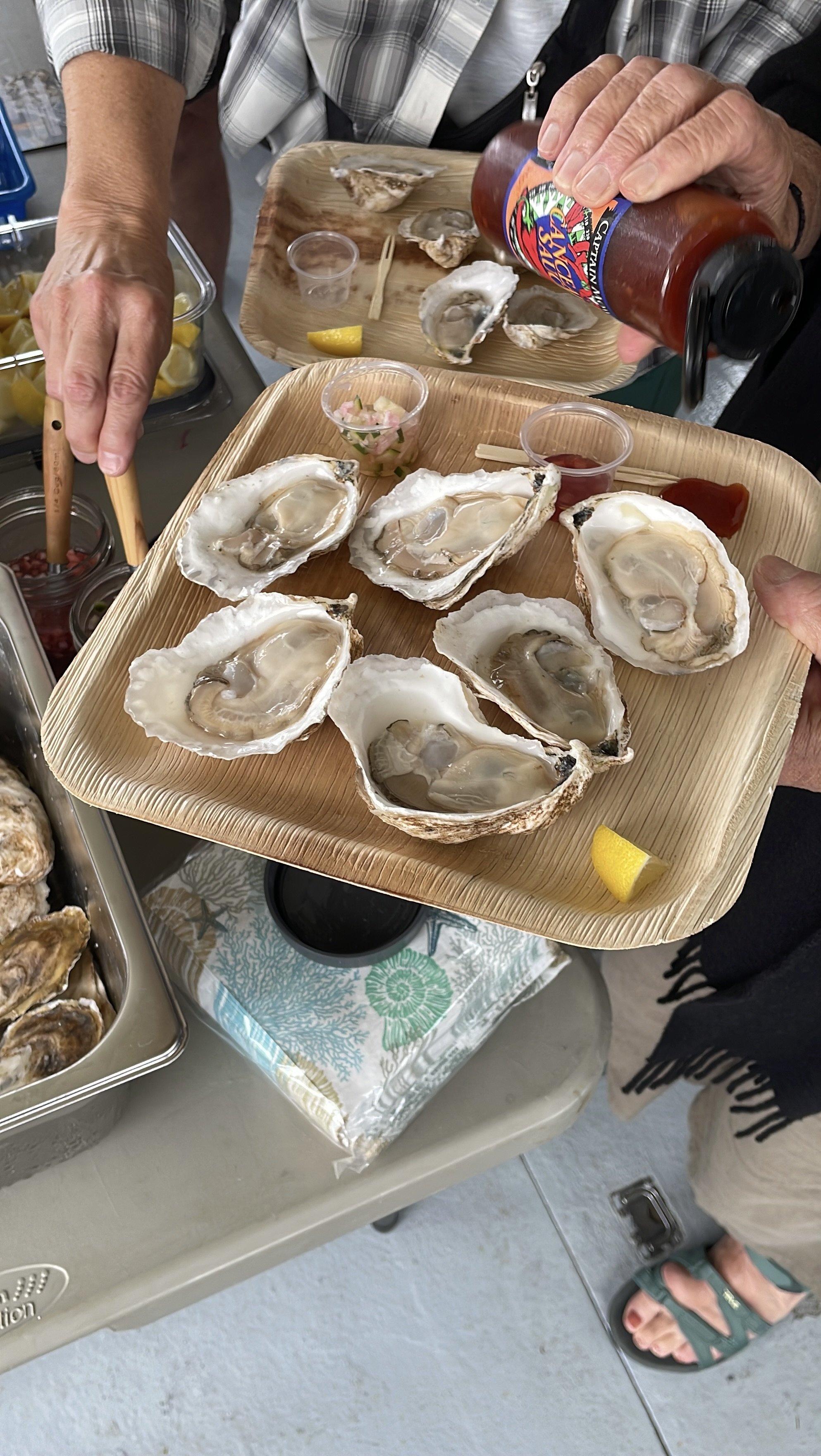 Person holding a tray of fresh oysters with lemon wedges and condiments, while another person is pouring hot sauce over oysters in the background.