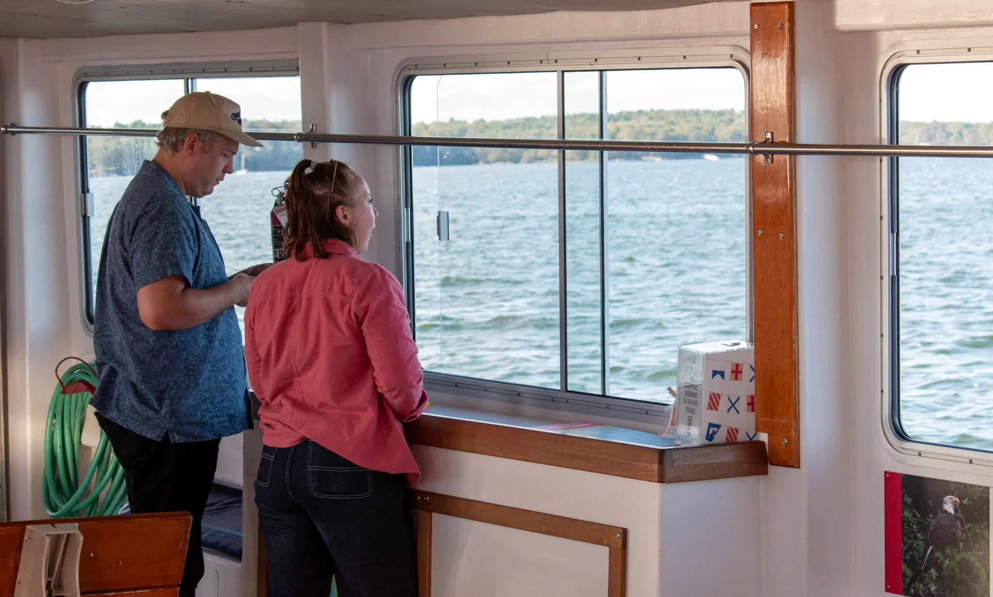 A man and woman standing inside a boat, looking out the window at a body of water with trees in the distance.